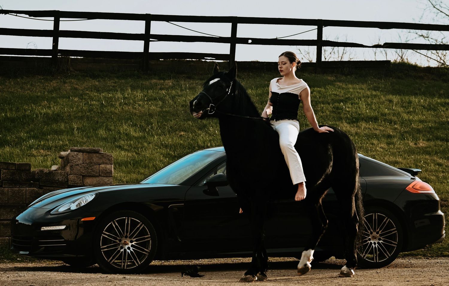Woman in white outfit riding a black horse rearing up beside a black Porsche sports car outdoors.