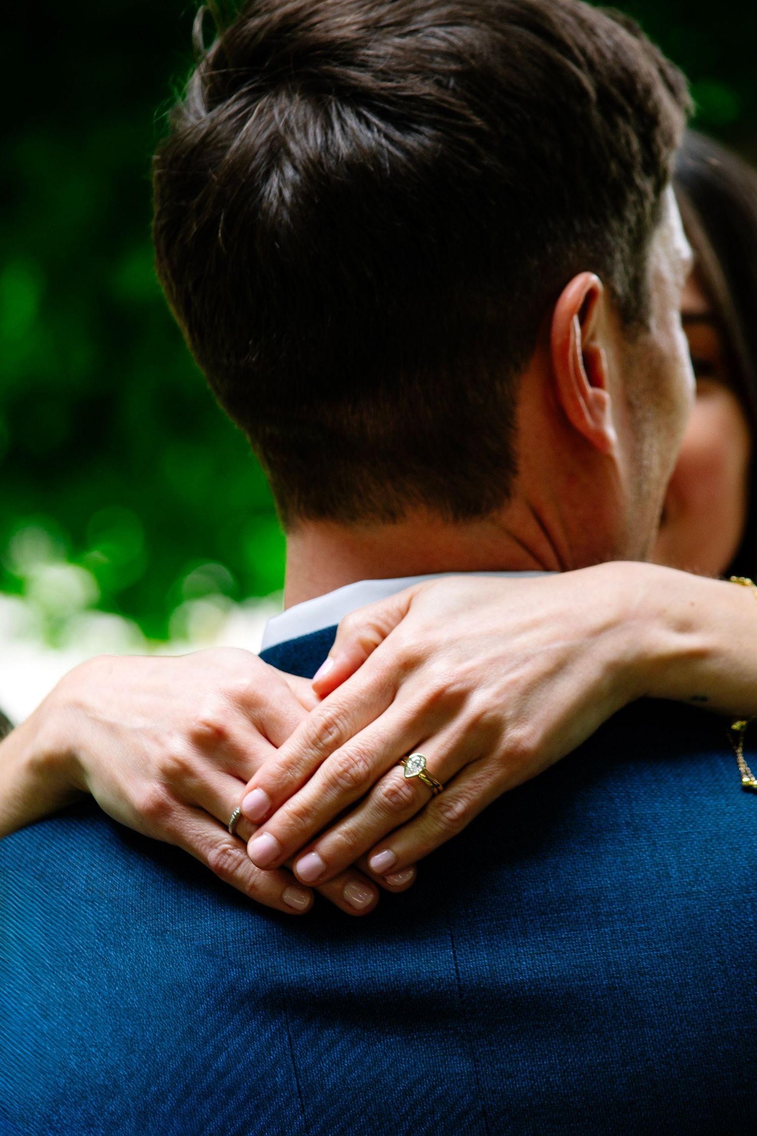 Close-up of bride's hand with diamond engagement ring resting on groom's navy suit shoulder during wedding.