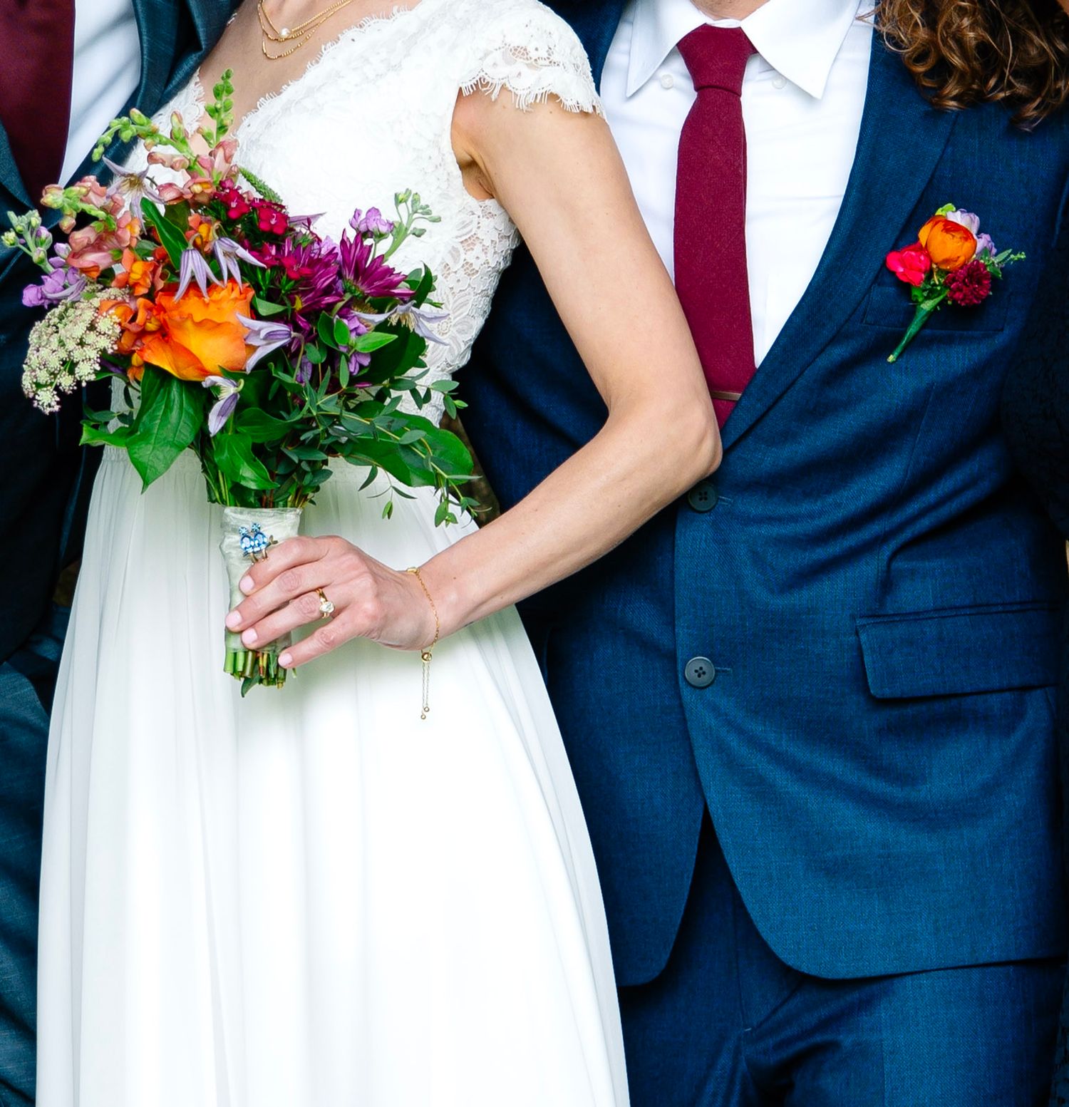 Bride in white lace dress holding colorful bouquet stands beside groom in navy suit with red tie and boutonniere.