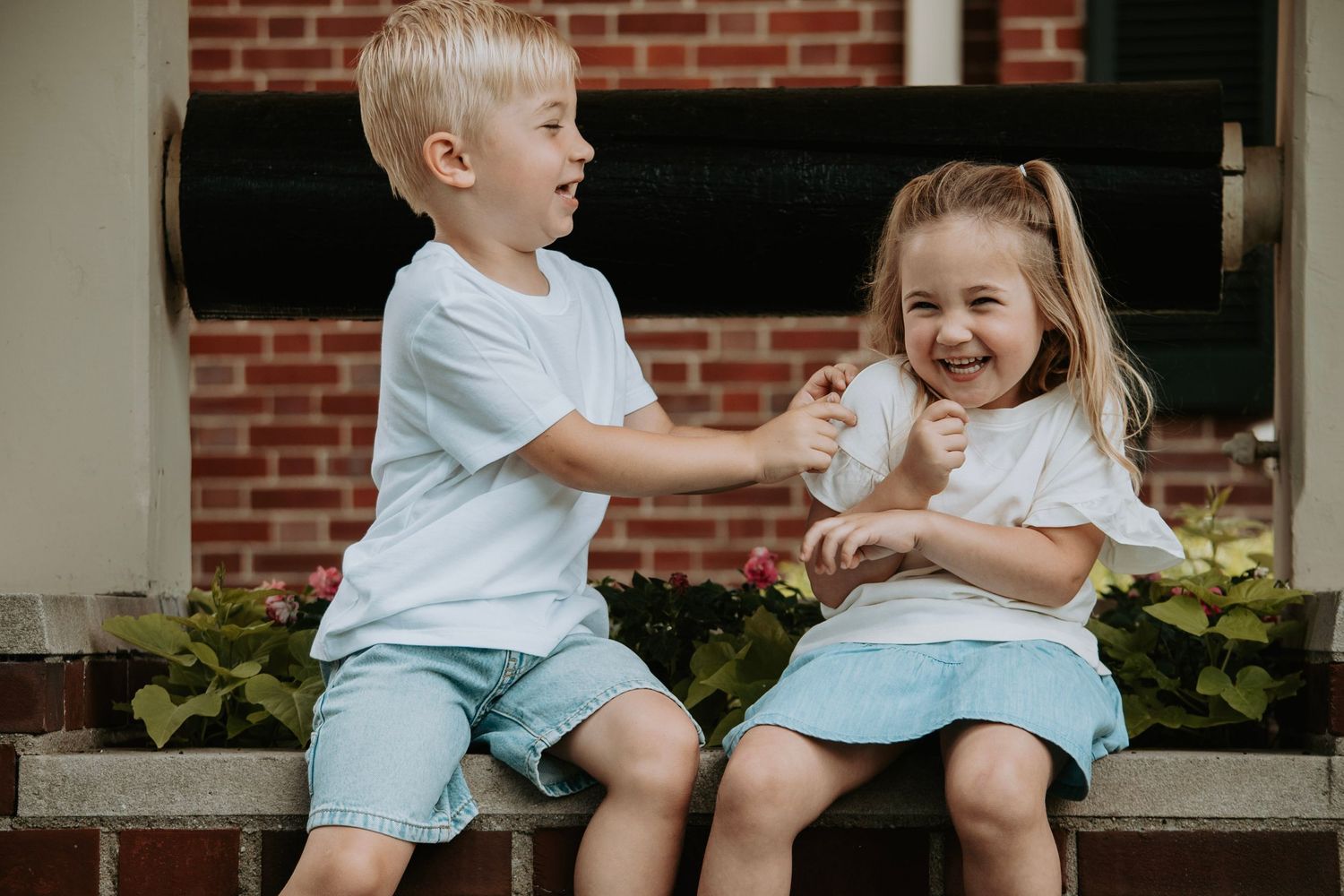 Two young children in matching white shirts and teal shorts sitting on steps, laughing and playing together outdoors.