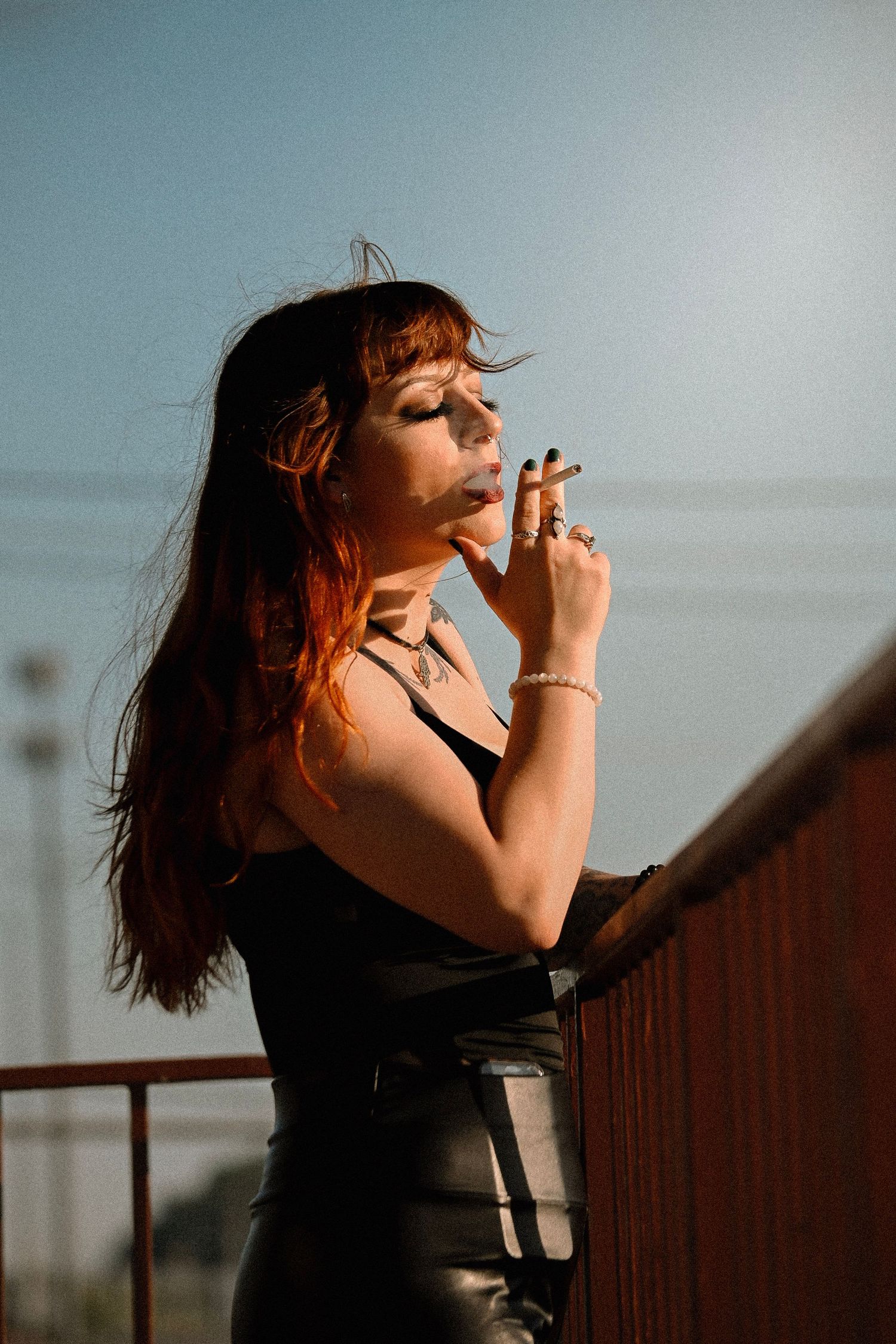 Young woman in black leather outfit smoking a cigar on a rooftop under a bright blue sky.