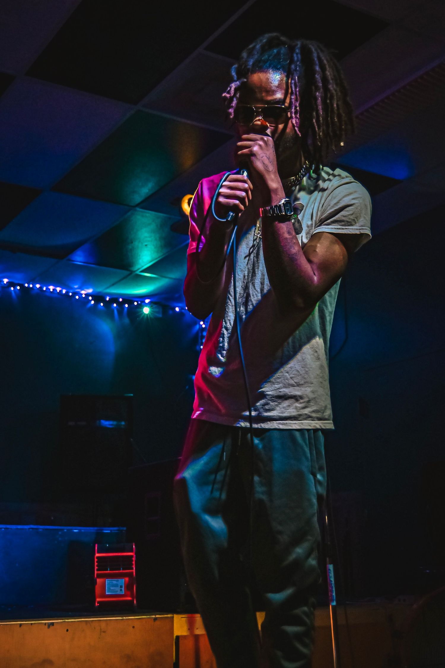 A performer with dreadlocks singing on stage illuminated by colorful stage lights.