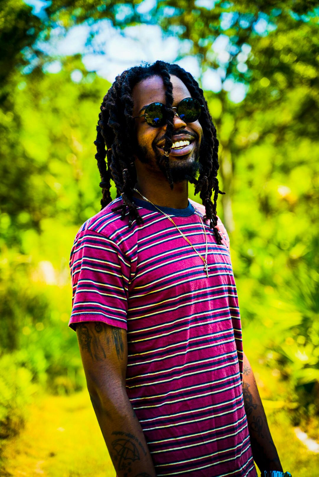 Person in striped pink shirt smiling outdoors with lush green foliage in background.