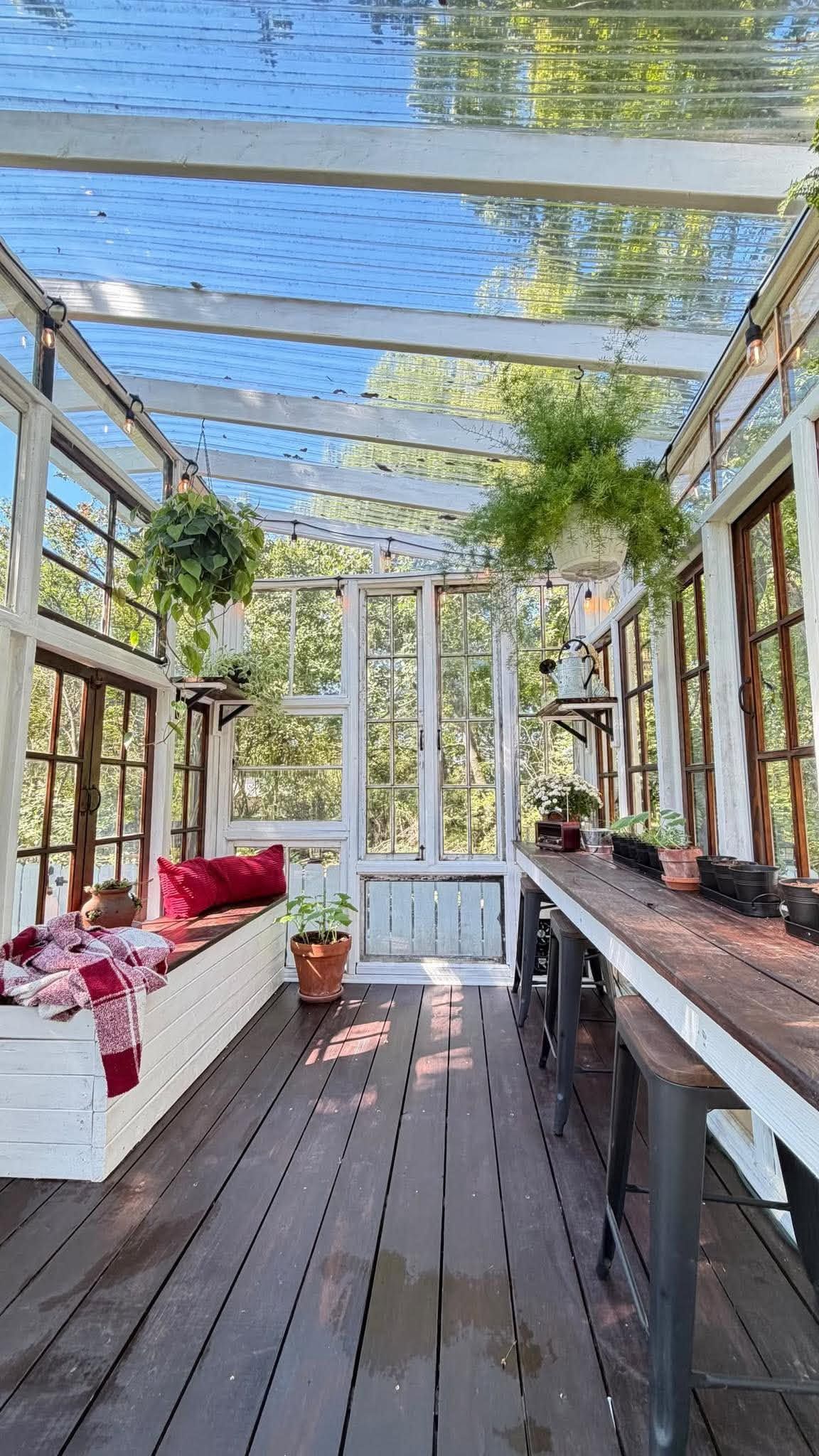 Bright sunroom with glass roof, hanging ferns, red cushion daybed, wooden floors, and potted plants on a rustic workbench.