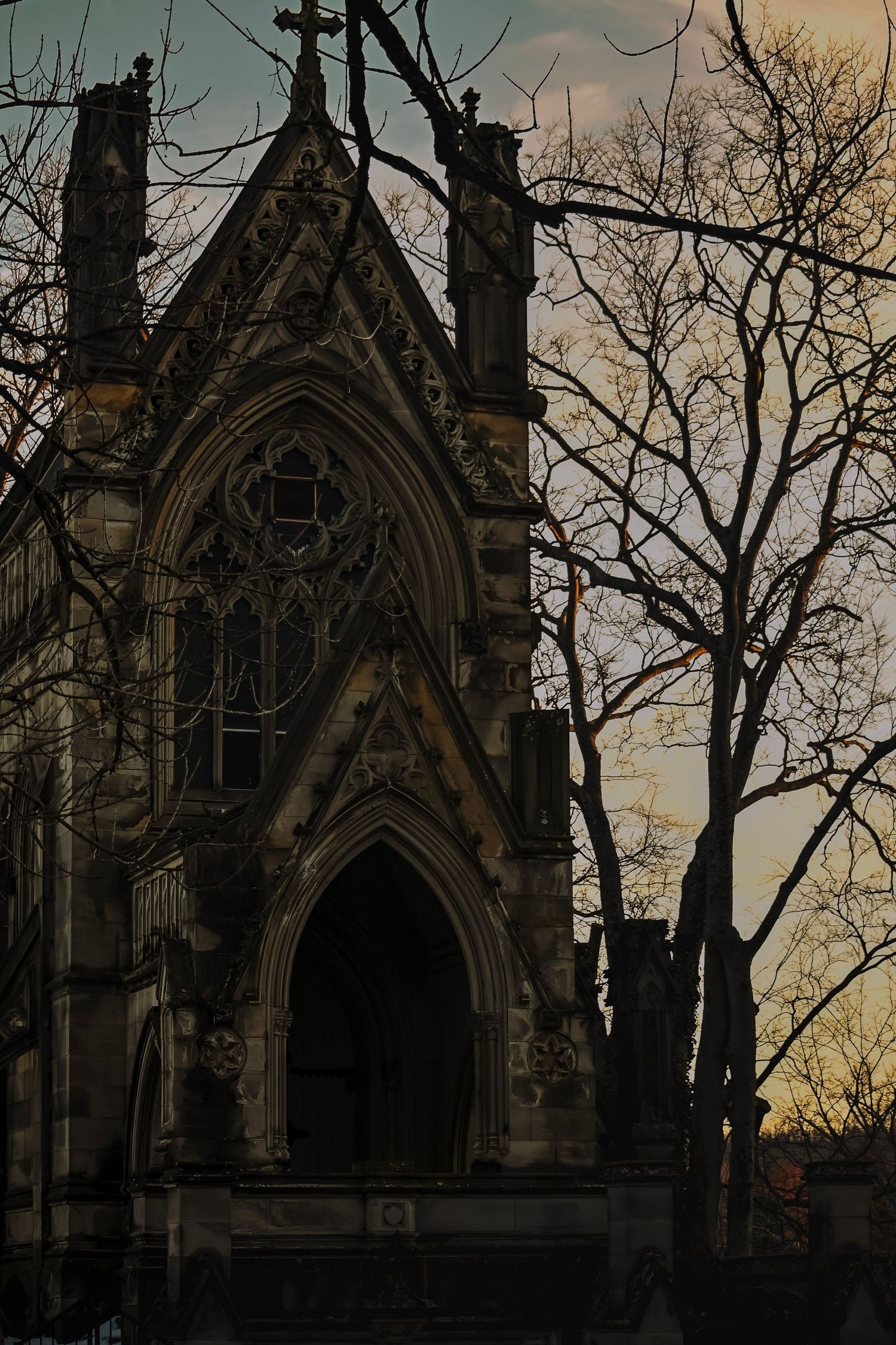 Gothic stone mausoleum silhouetted against a moody sunset sky with bare winter trees in the foreground.