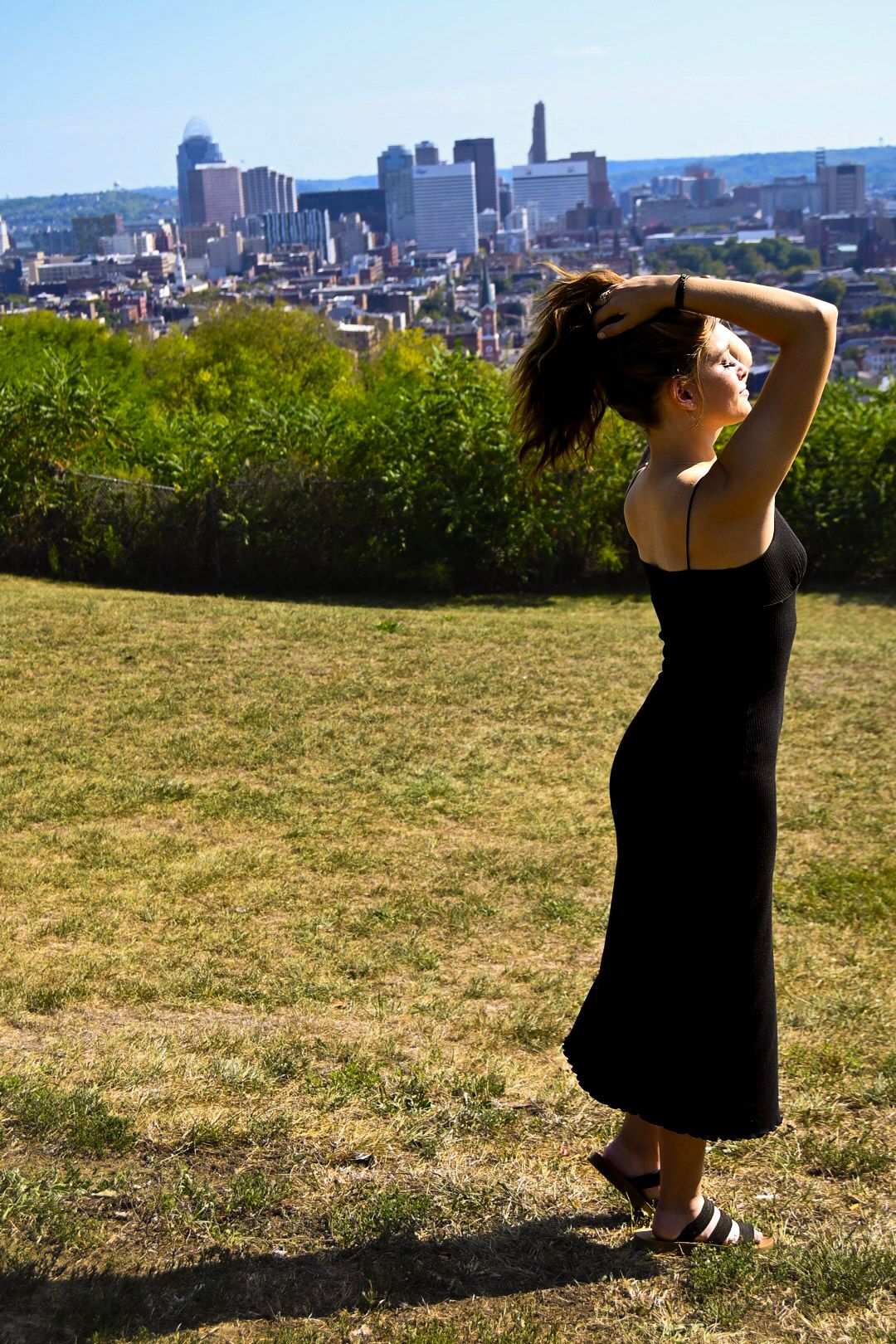 A silhouetted figure in a black dress stands on a hilltop overlooking the Cincinnati skyline on a sunny day.