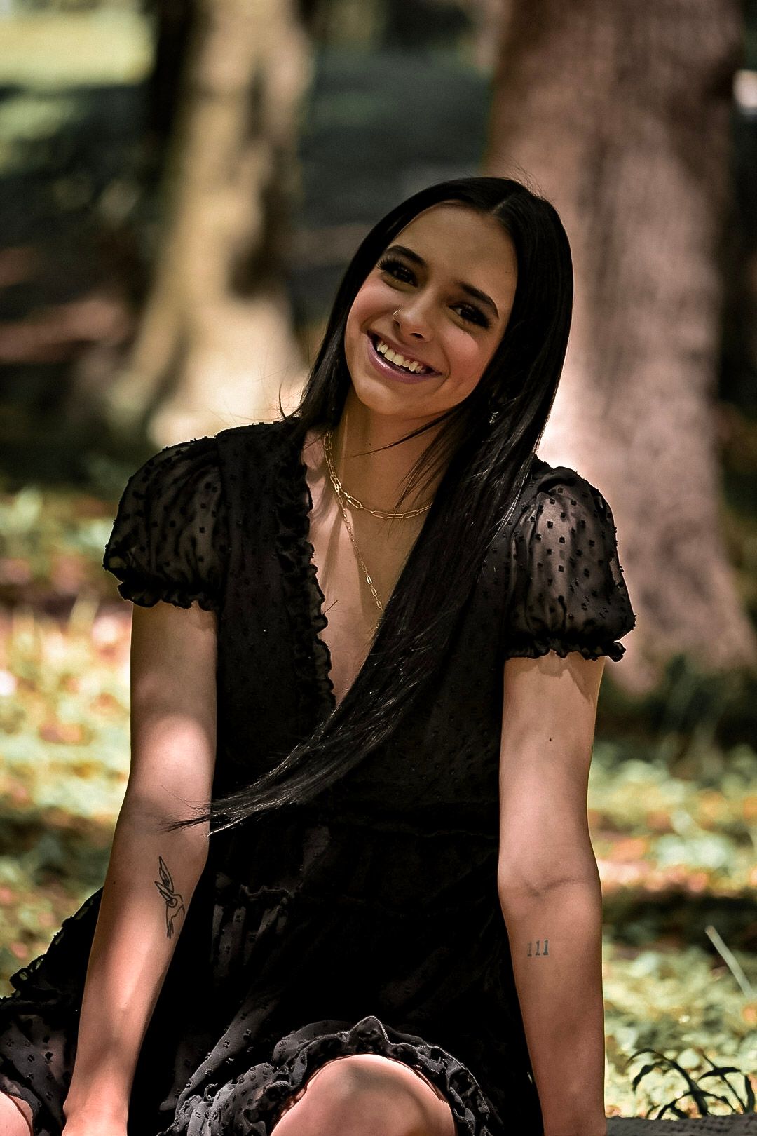 Person in black lace dress smiling while sitting outdoors among autumn leaves.
