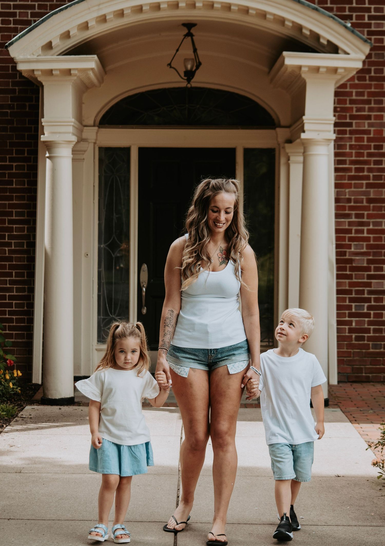 Smiling mom holds hands with her two young children in coordinating outfits in front of a brick home entrance.