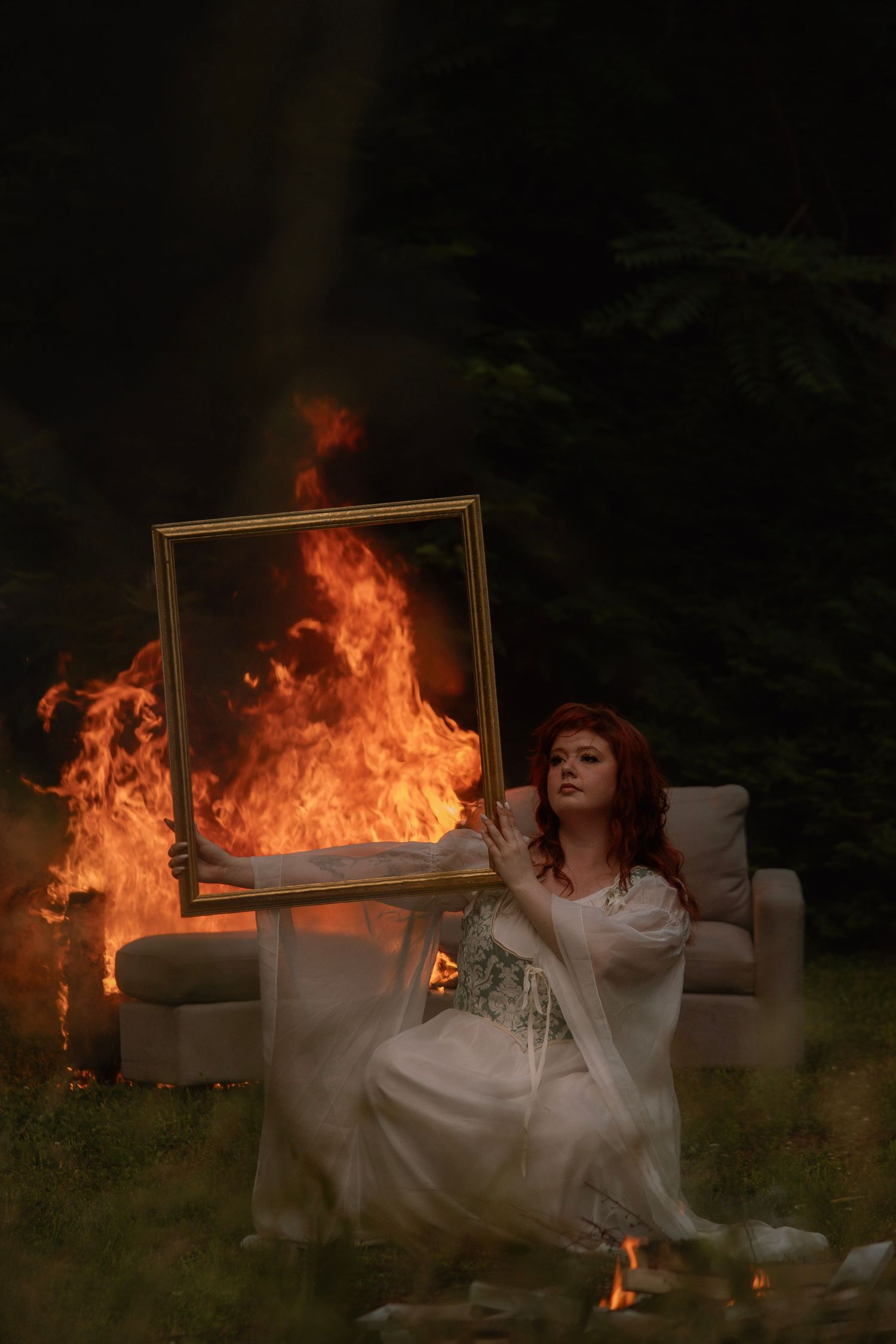 A red-haired woman in a white gown holds a burning gold frame while sitting outdoors at night.