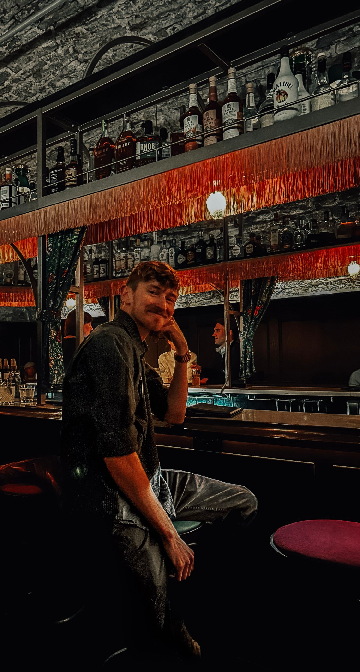 Moody bar interior with red lighting featuring bottles on shelves and person at counter.