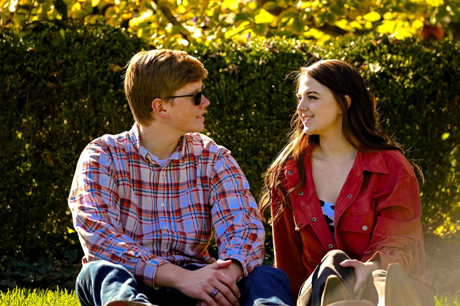 A couple in plaid and red jackets sitting together outdoors during golden hour sunlight in a park setting.
