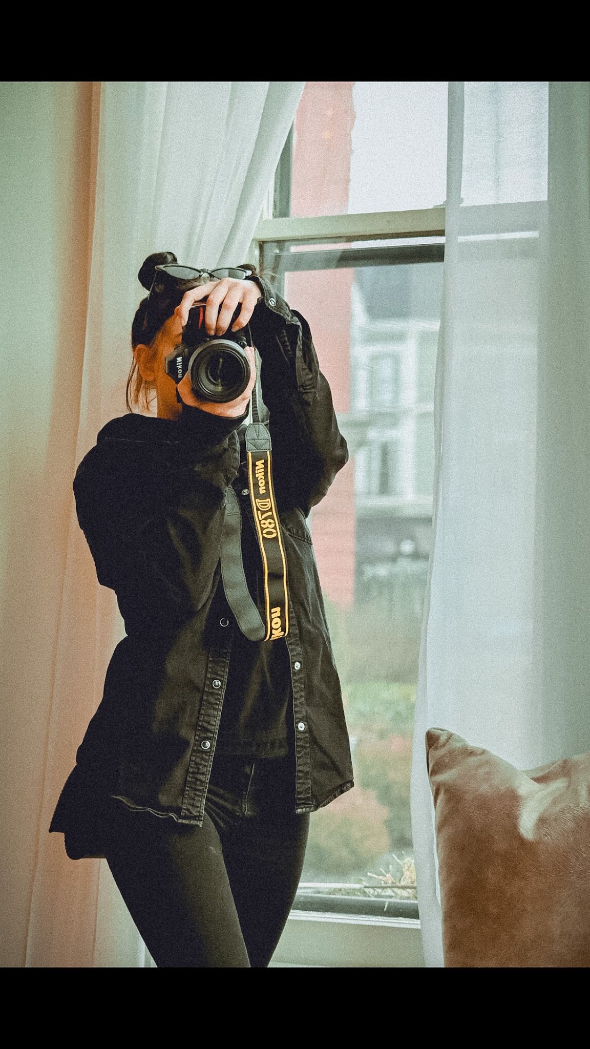 A photographer in dark clothing holds a camera near a window with soft natural light filtering through sheer curtains.