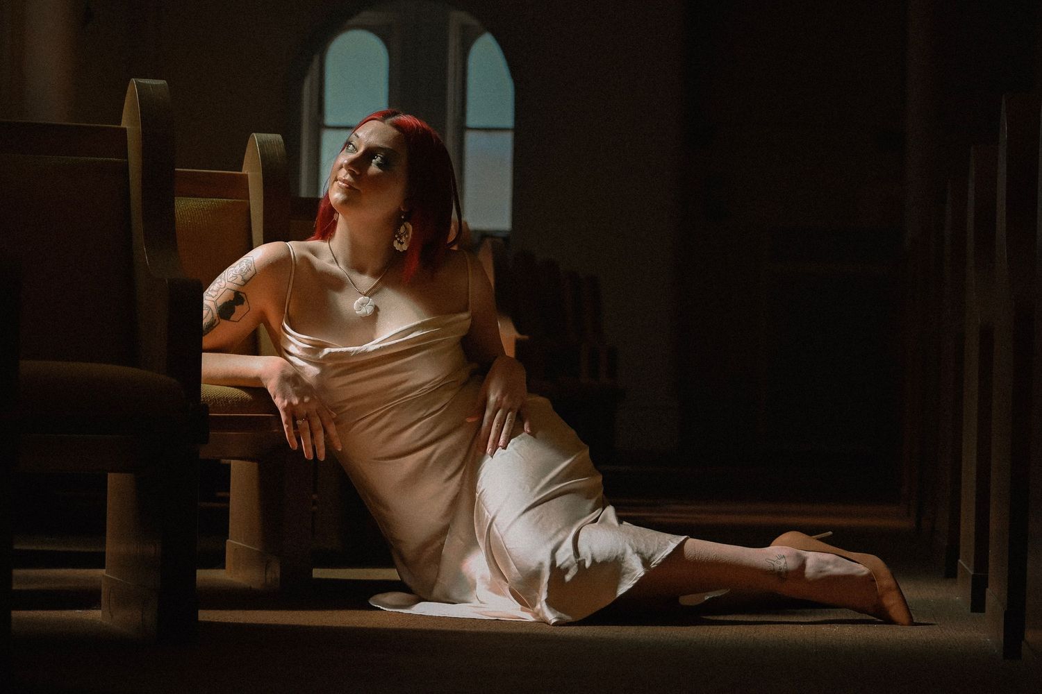 Red-haired woman in white slip dress sitting gracefully on church floor, bathed in dramatic light.