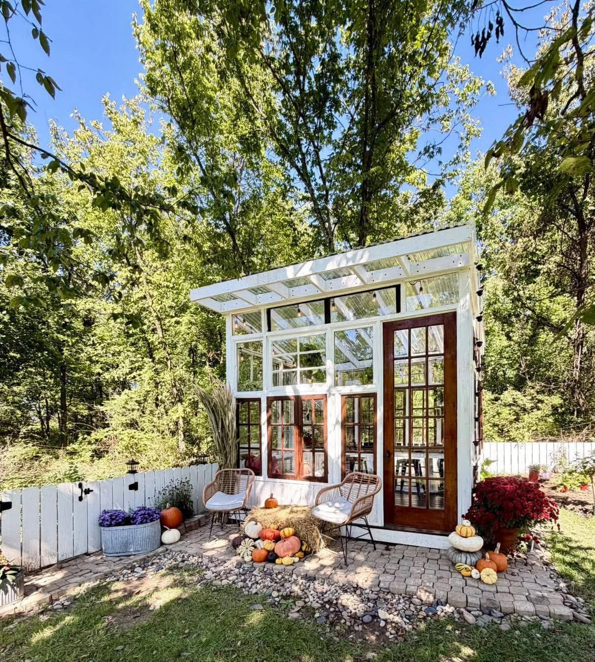 Charming glass greenhouse with white frame, French doors, pumpkins, and autumn decor surrounded by lush green trees in a backyard.