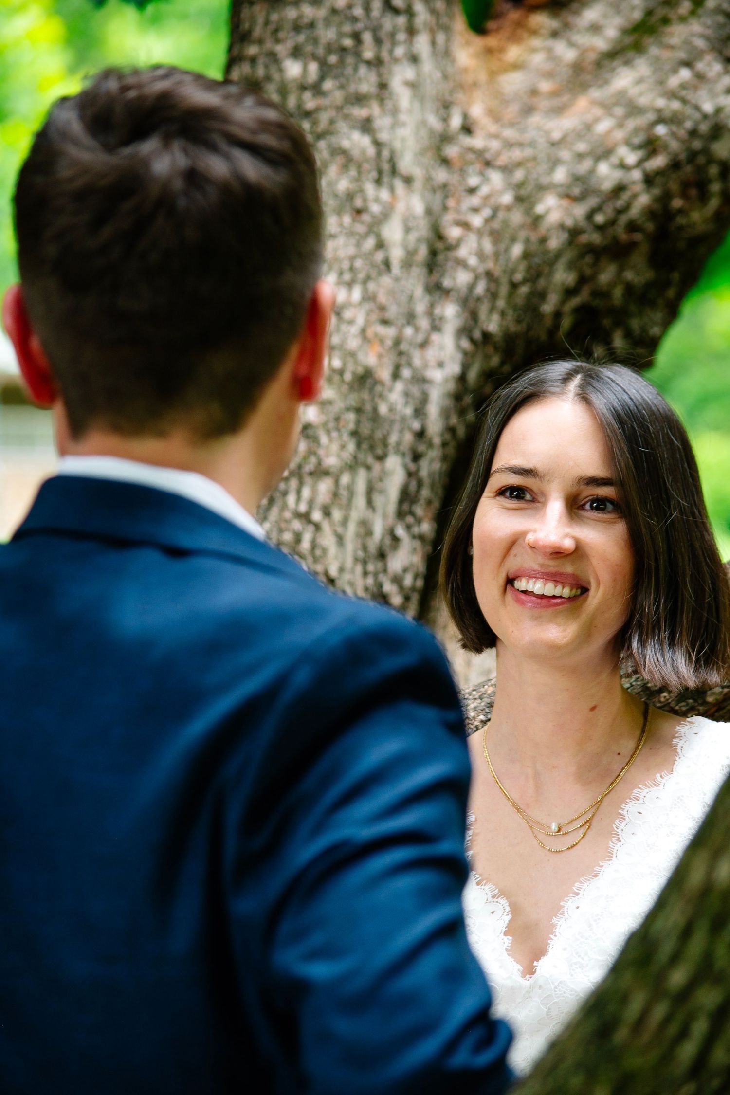 Smiling bride in white lace dress gazes at groom in navy suit near a large tree during outdoor wedding.