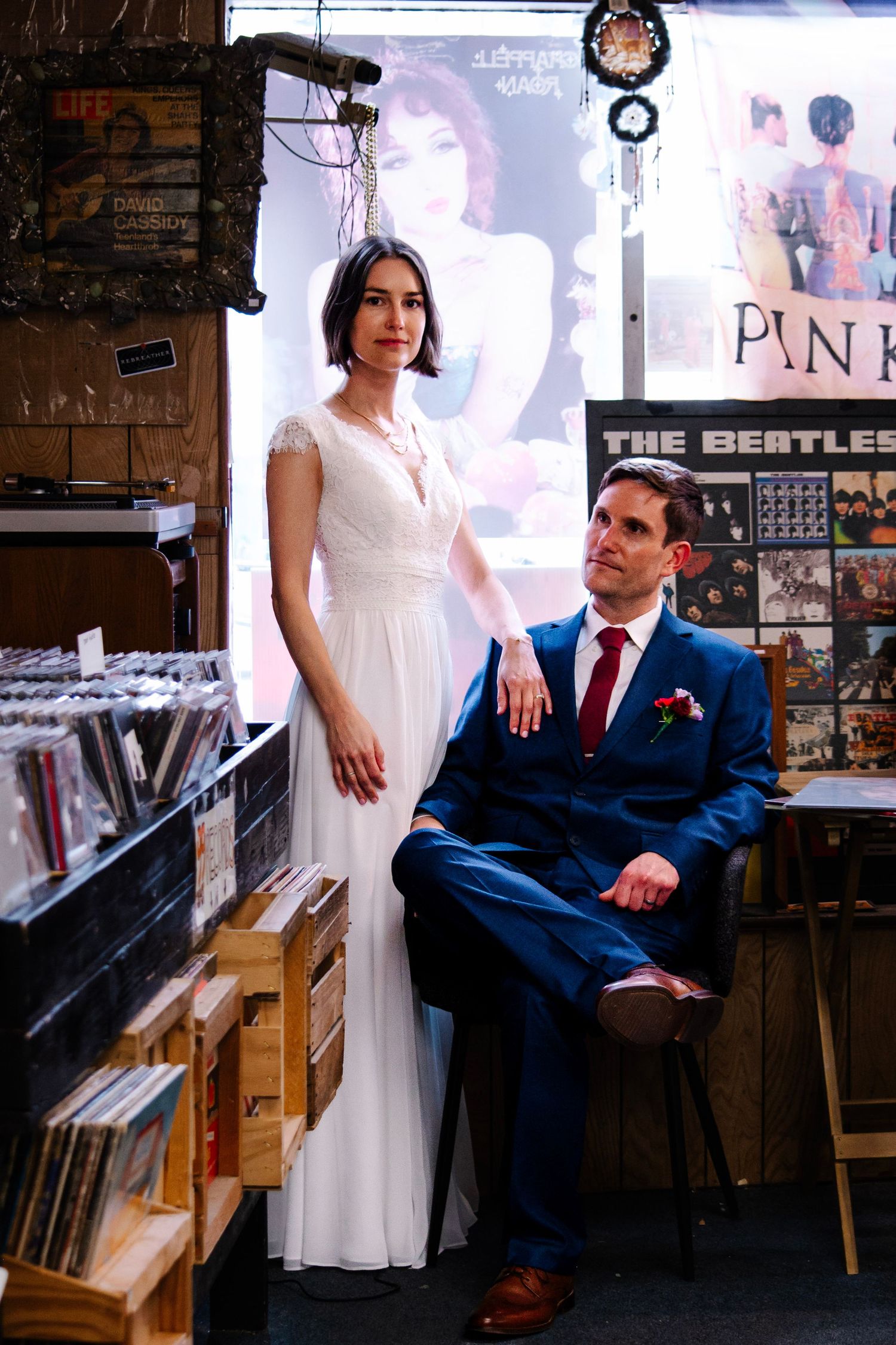 Bride and groom posing in a vintage record store, surrounded by vinyl albums including The Beatles poster.