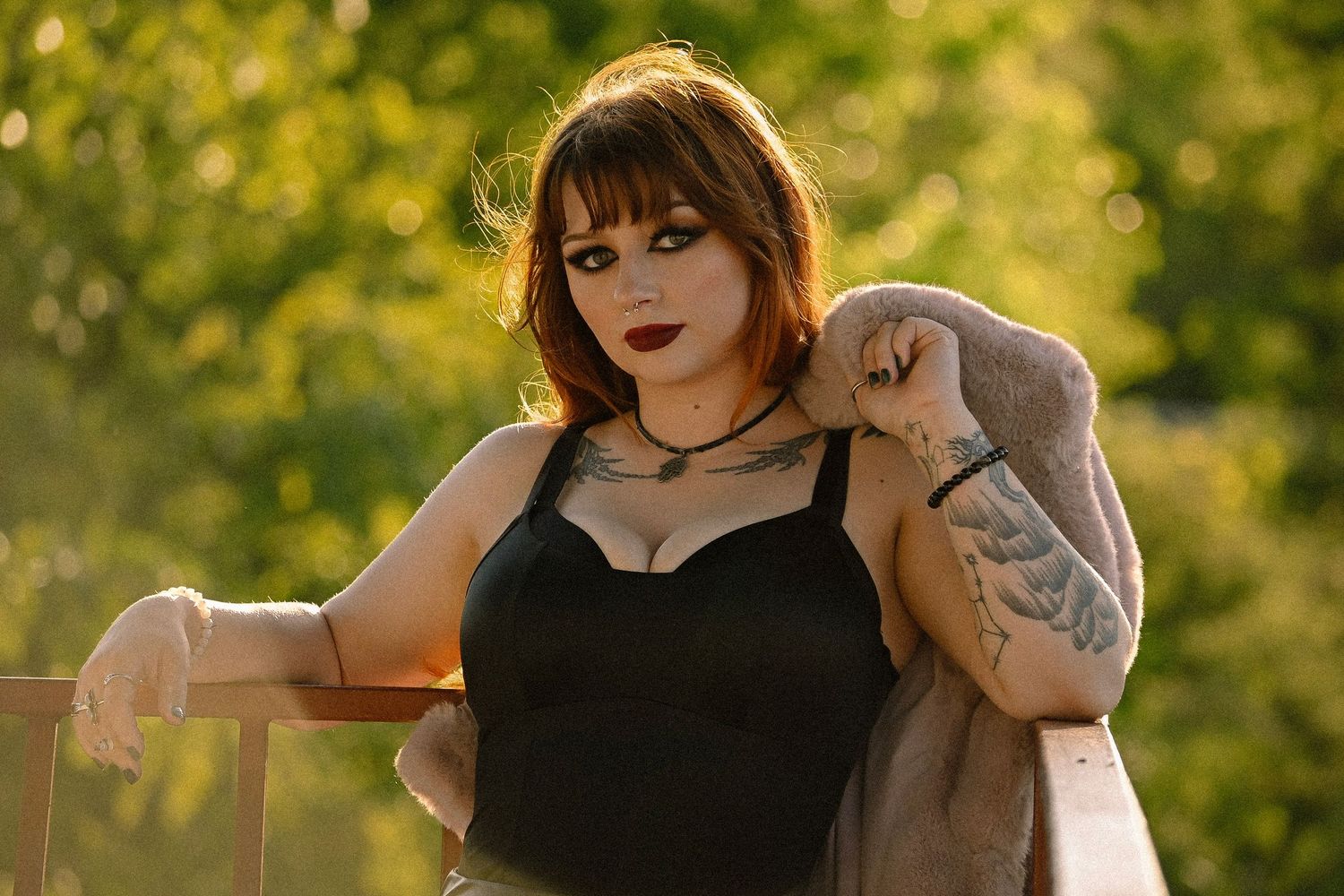 A tattooed woman in a black top and pink jacket poses outdoors with lush green trees in golden sunlight.