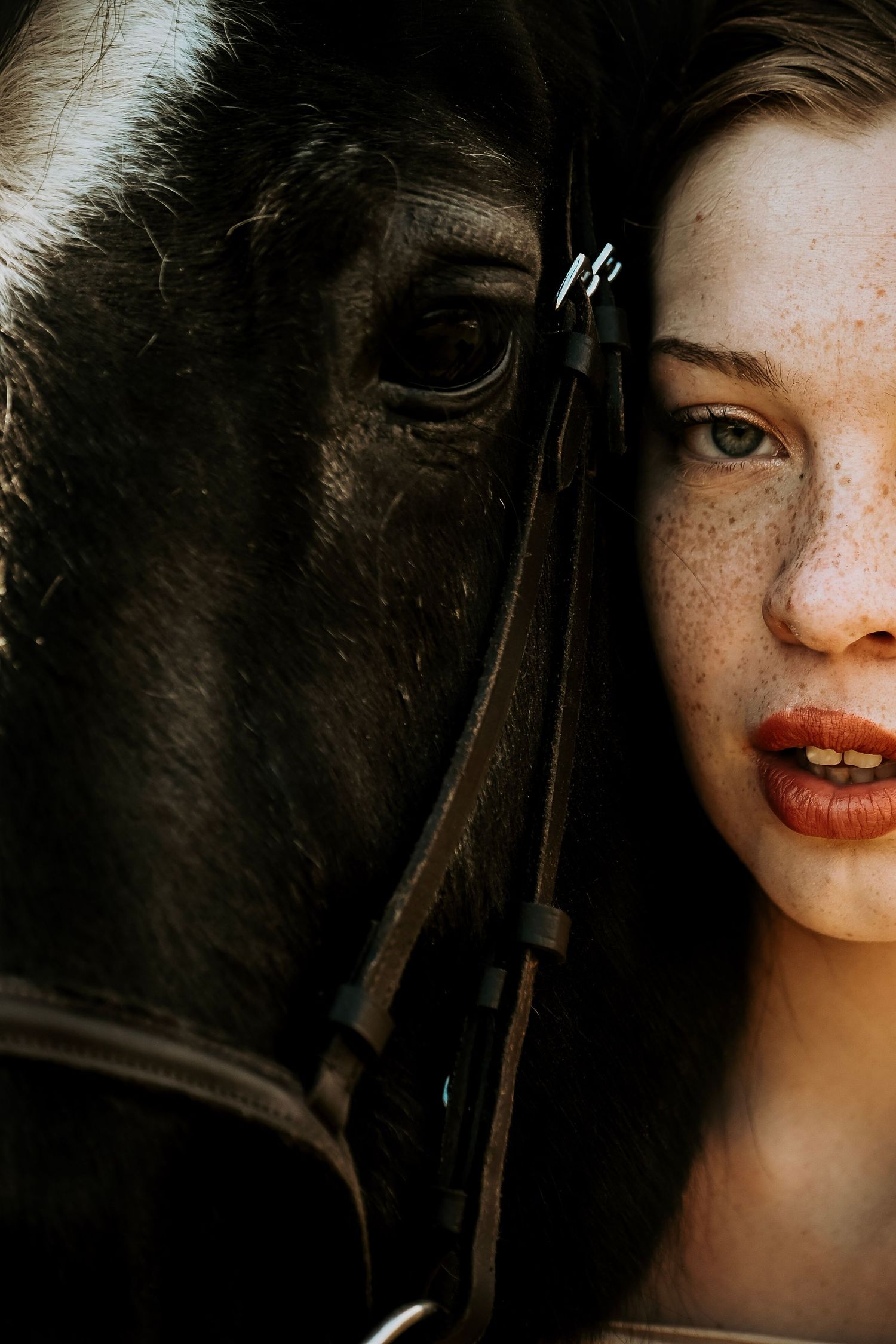 Close-up portrait showing a black horse and person with freckles and red lipstick in dramatic lighting.