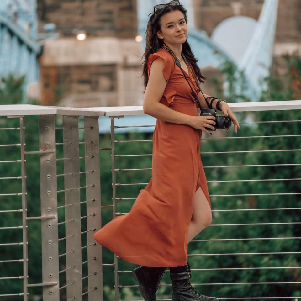 A photographer in a rust-colored dress and boots stands on a wooden deck overlooking a scenic outdoor view.