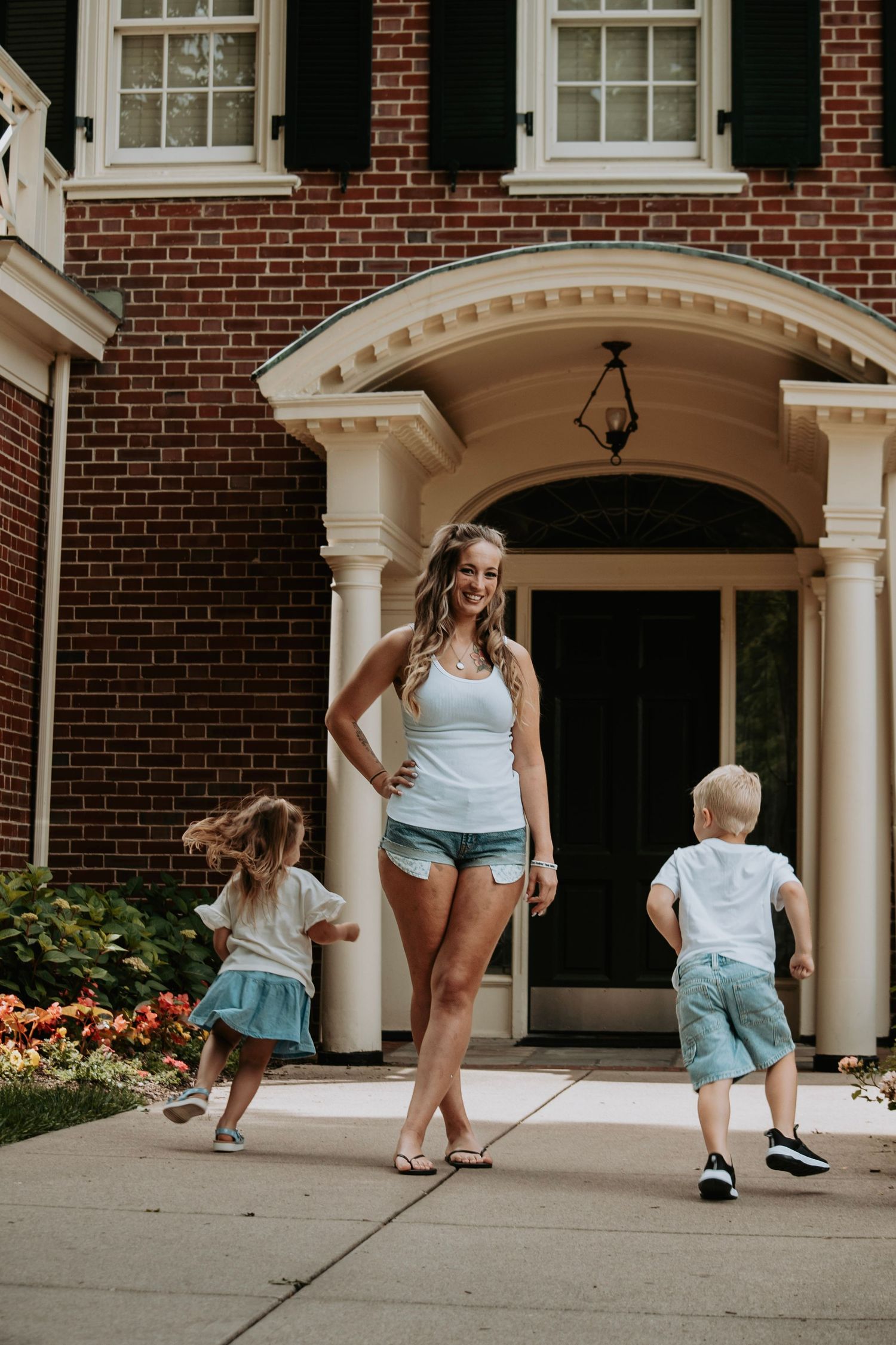 A smiling woman in denim shorts stands with two young children on a driveway in front of a brick colonial home.