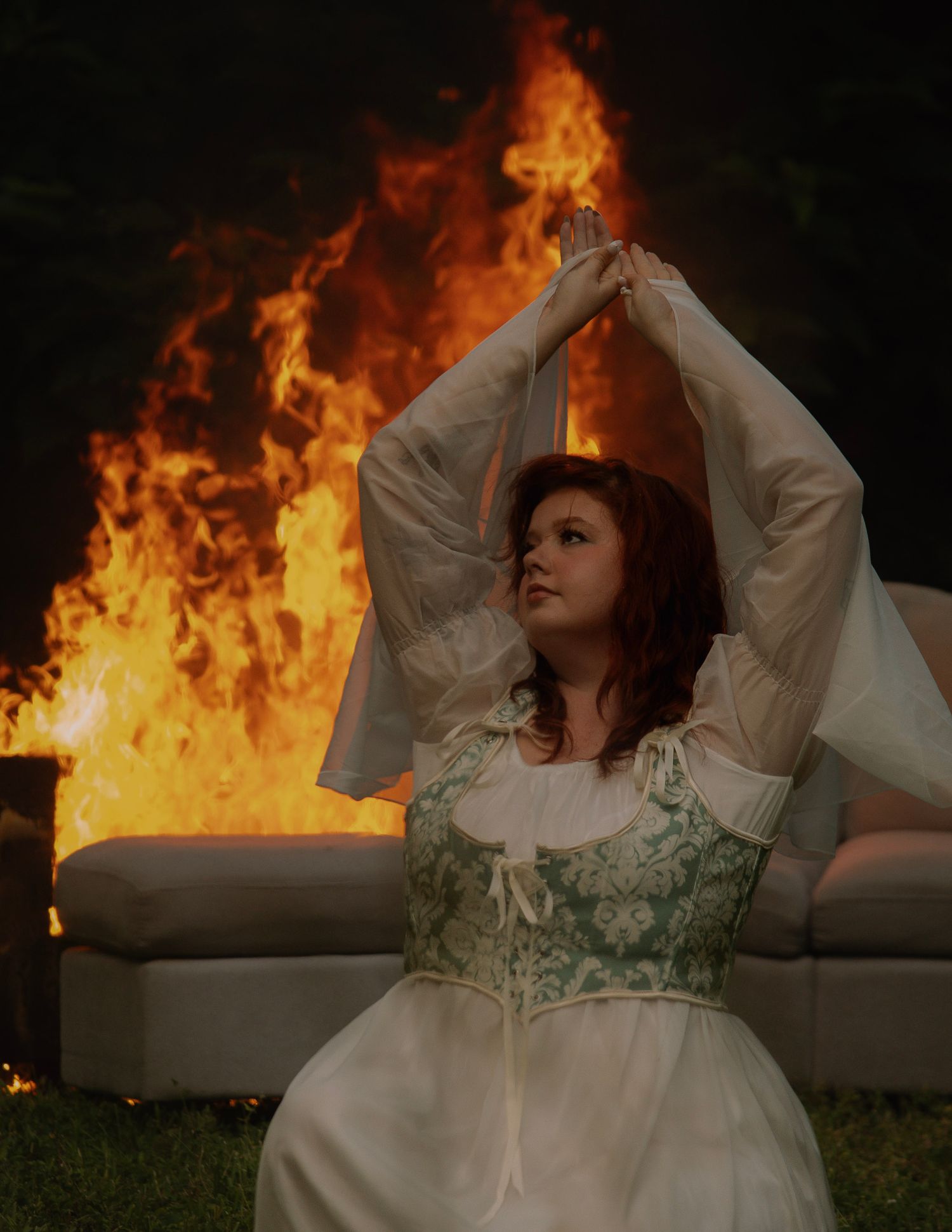 Woman in white and green corset dress poses dramatically with arms raised before a large outdoor fire at night.