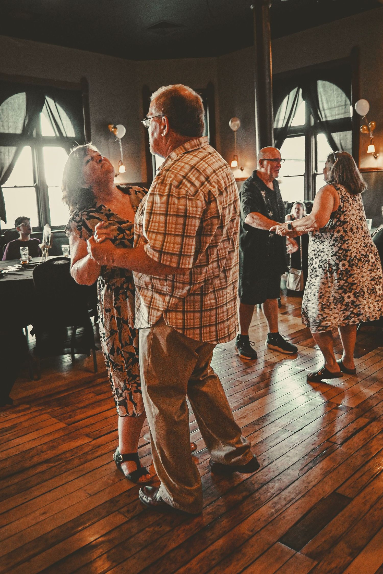 Couples dancing together on a wooden floor in a vintage-style room with arched windows.