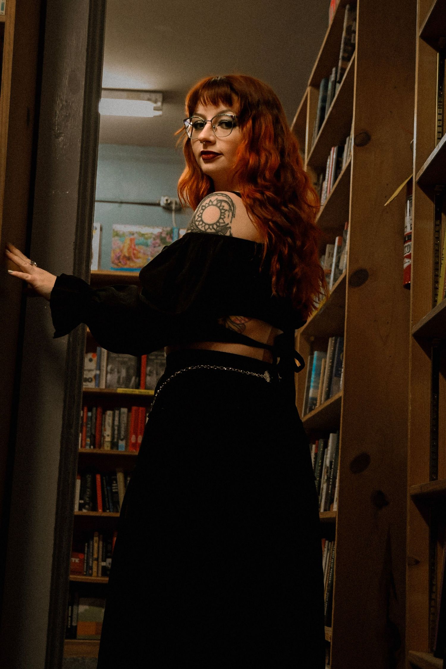 Woman with red hair in a black dress poses dramatically between library bookshelves in a dark moody setting.