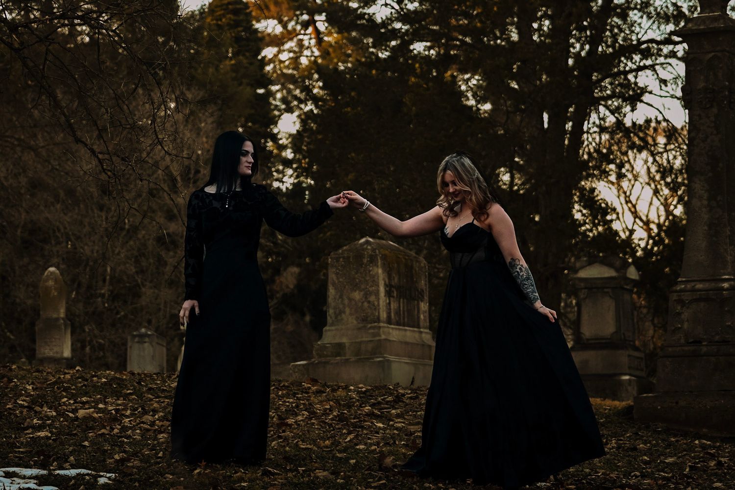 Two figures in black dresses dance together among fallen autumn leaves in a moody cemetery setting at dusk.