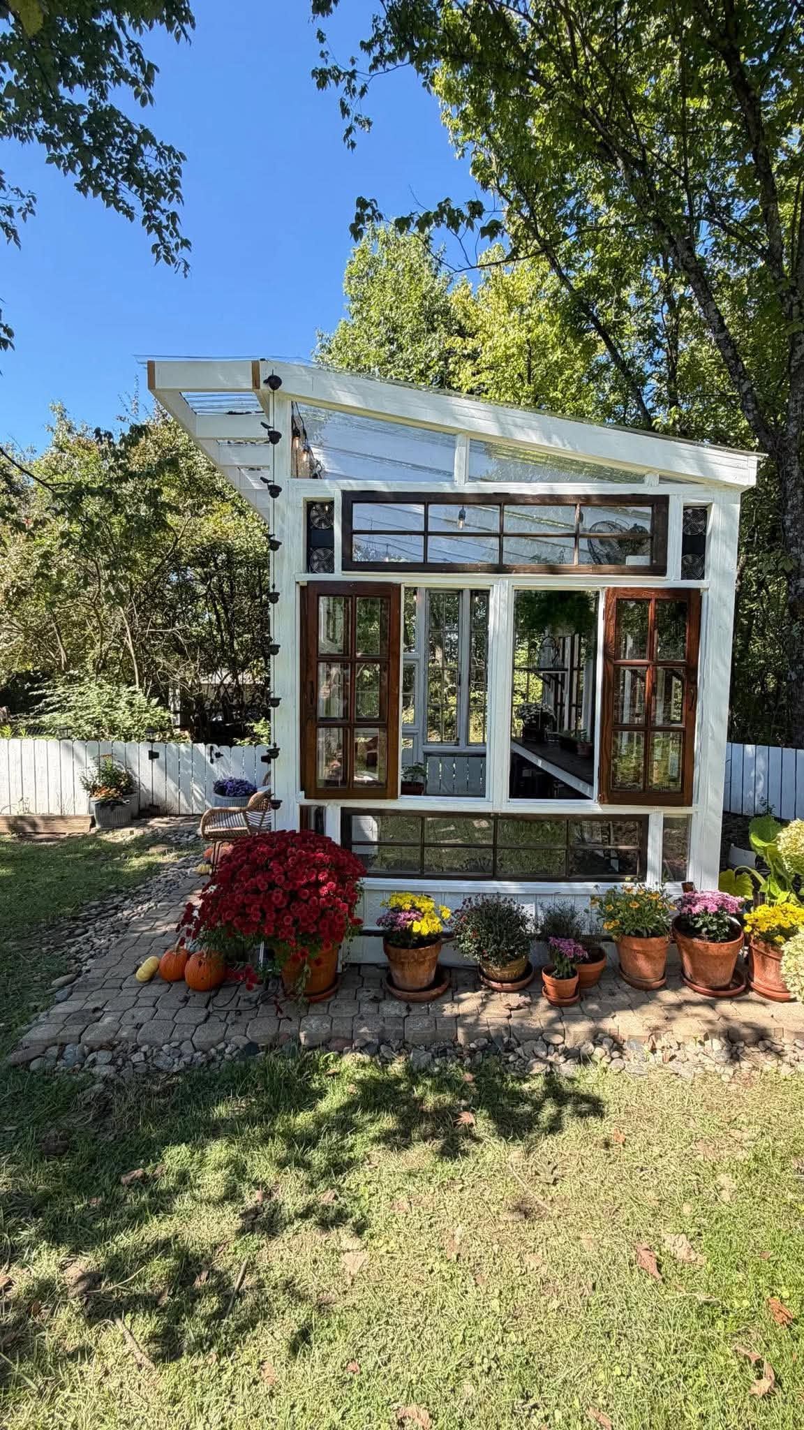 Charming DIY greenhouse built from vintage windows and doors, surrounded by colorful potted flowers and pumpkins in a backyard garden.