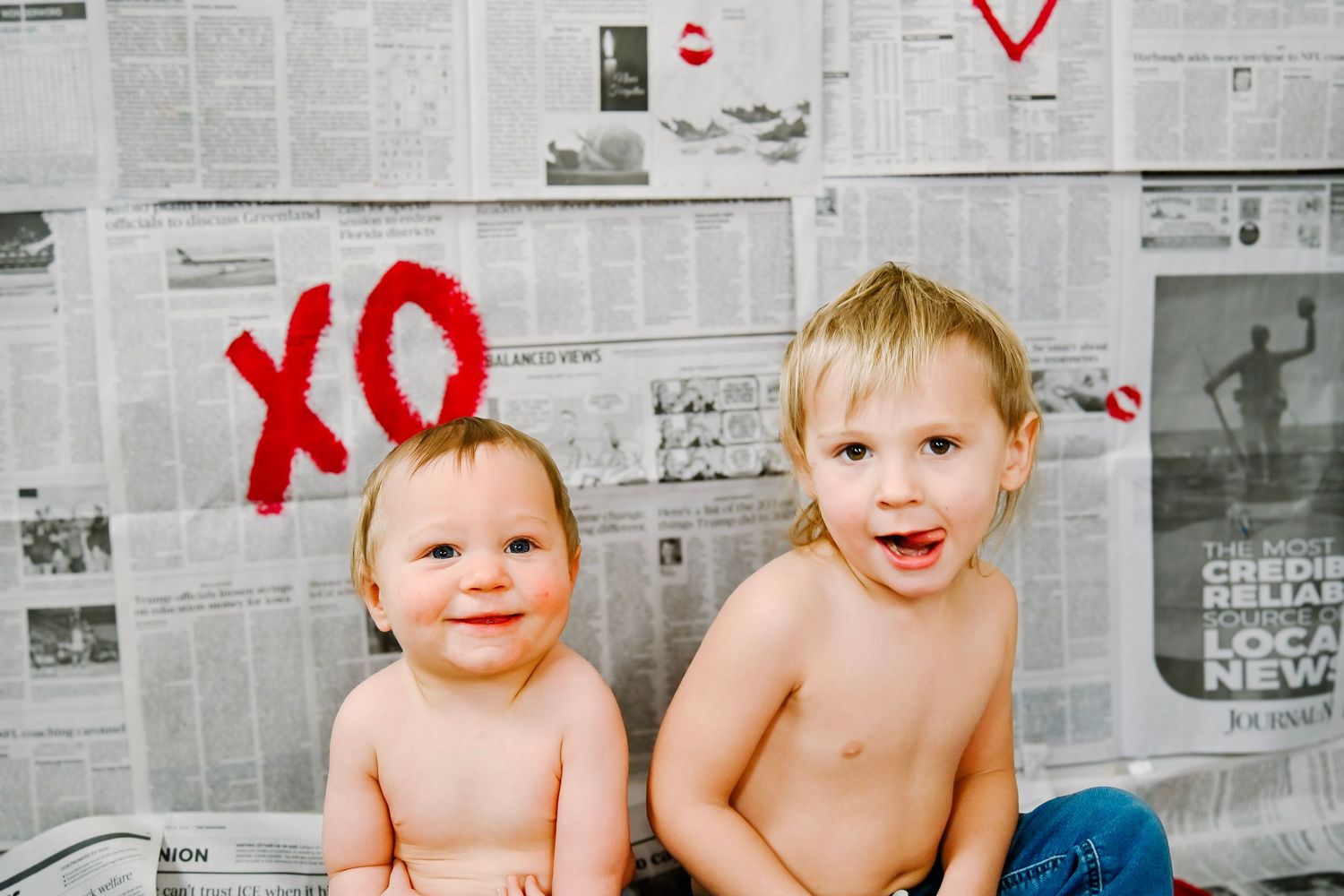 Babies sit against a newspaper wall backdrop while covered in red paint during a creative photo session.