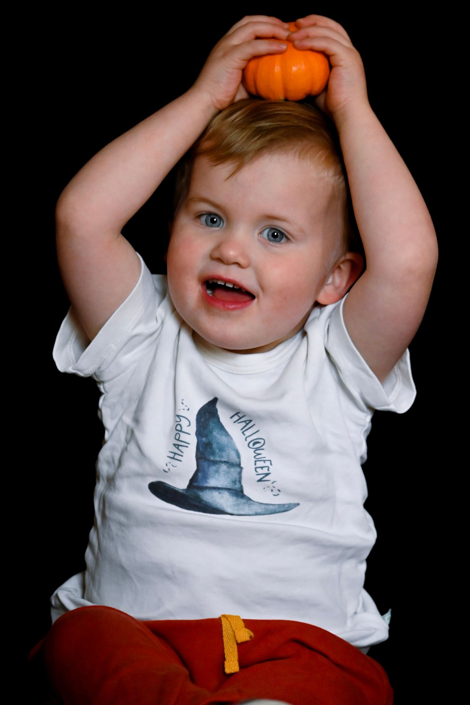 Smiling child in white t-shirt holds small orange pumpkin on head against black background.