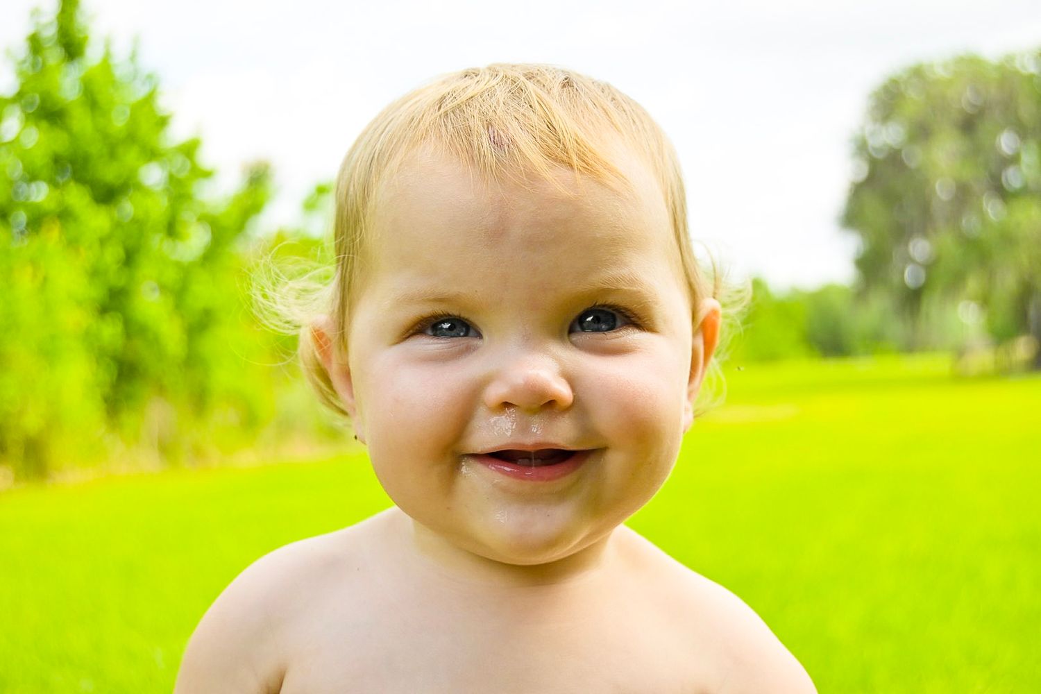 Baby with bright eyes and rosy cheeks smiling in a sunny green field.