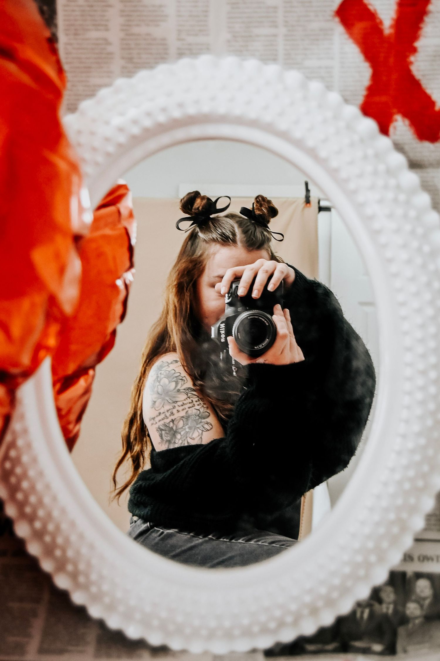 Mirror reflection shows photographer with camera taking self portrait in ornate white oval frame with red accents.
