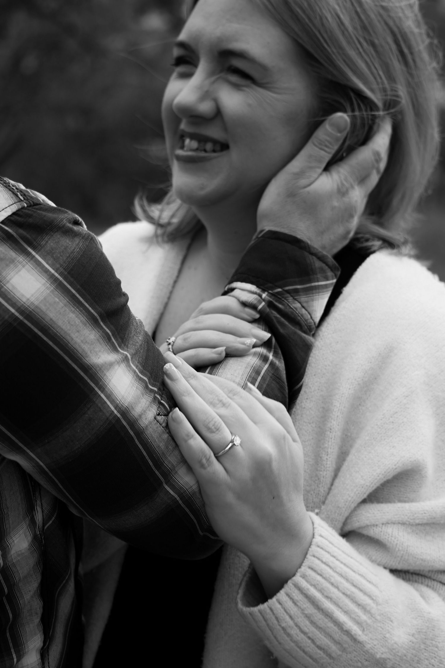 Black and white photo of a smiling woman in a cardigan holding a plaid jacket, wearing a ring outdoors.