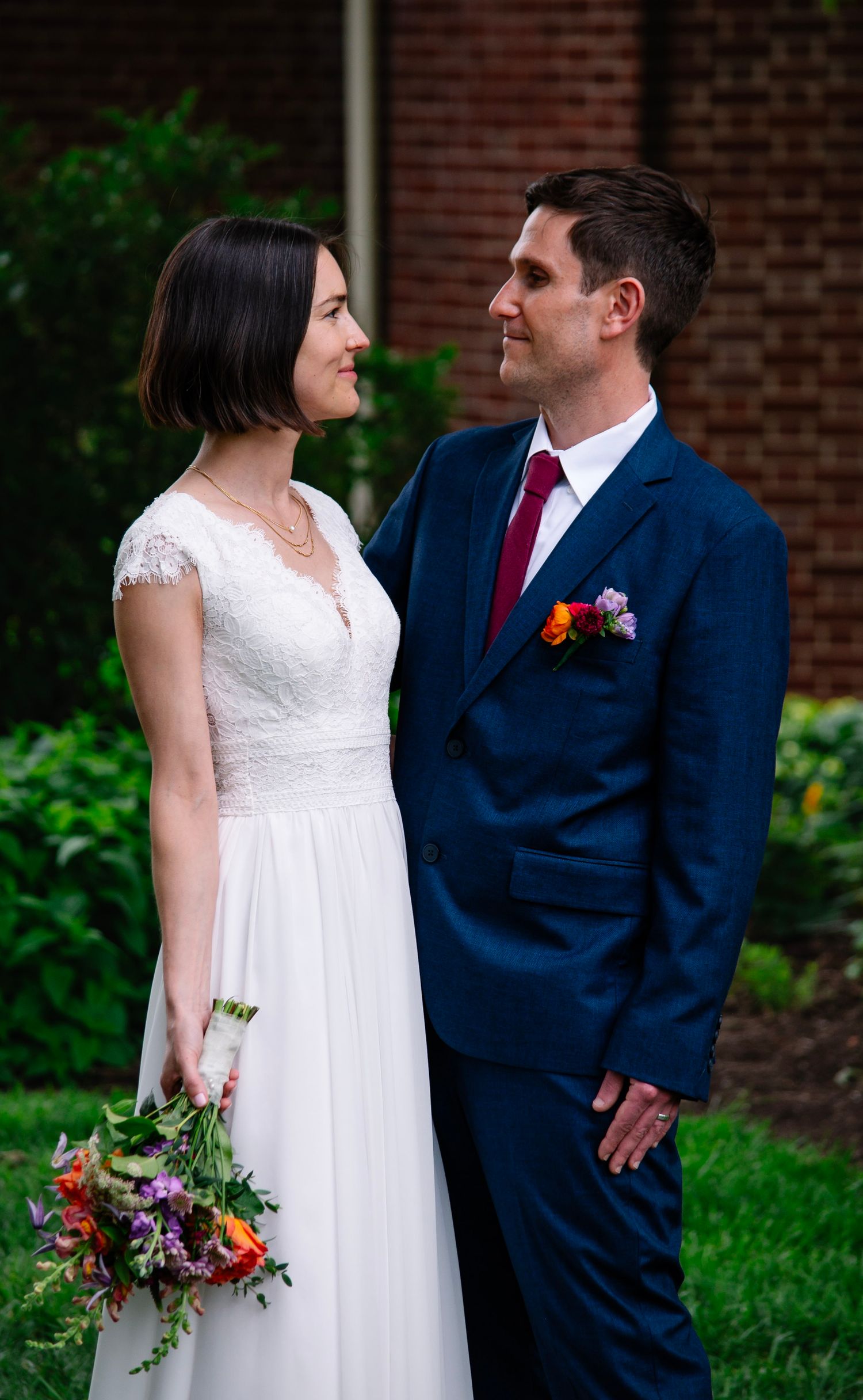 A bride in a white lace gown holds colorful flowers as she and her groom in a navy suit share a loving gaze outdoors.