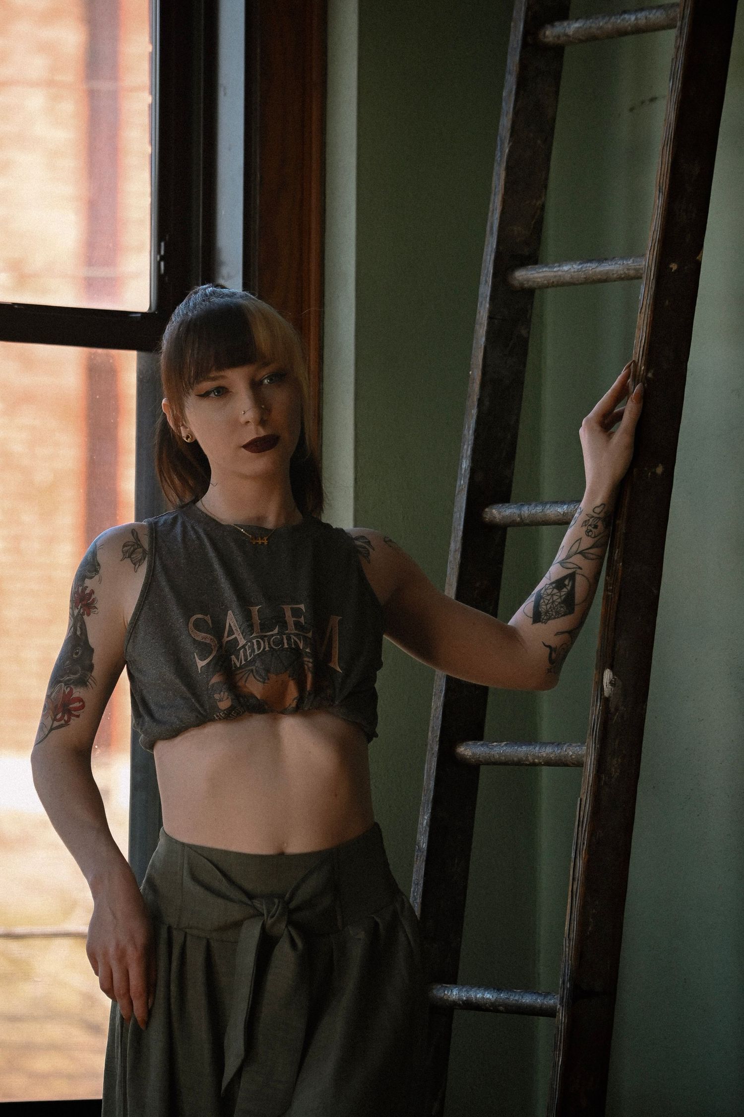 Moody portrait showing a cropped top and olive pants against a window with a vintage ladder.