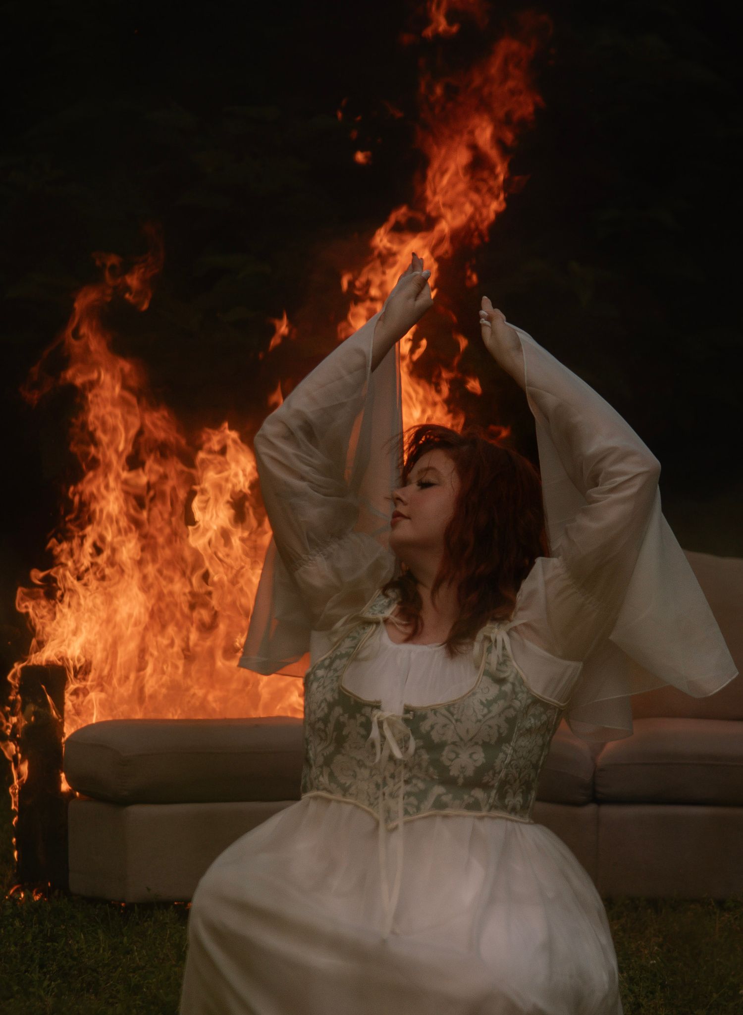 Woman in medieval dress sits serenely before dramatic flames engulfing a sofa outdoors at night.