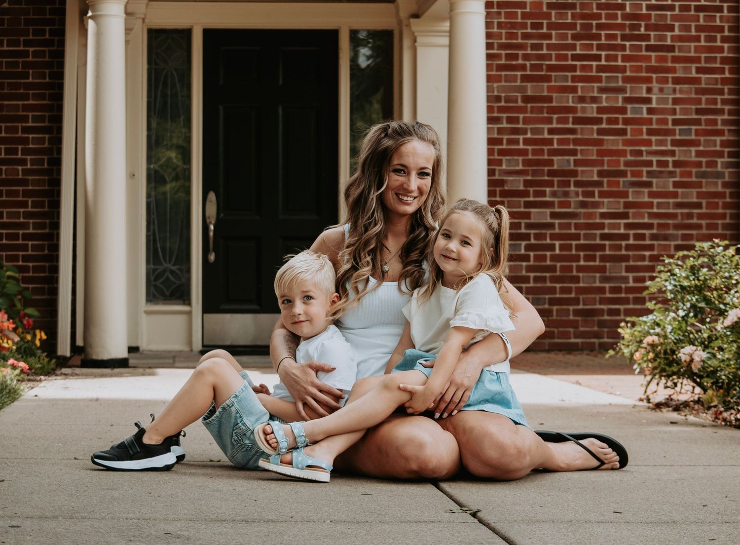 Happy mom sits on a driveway with her two young children in coordinating white and blue outfits by a brick house.