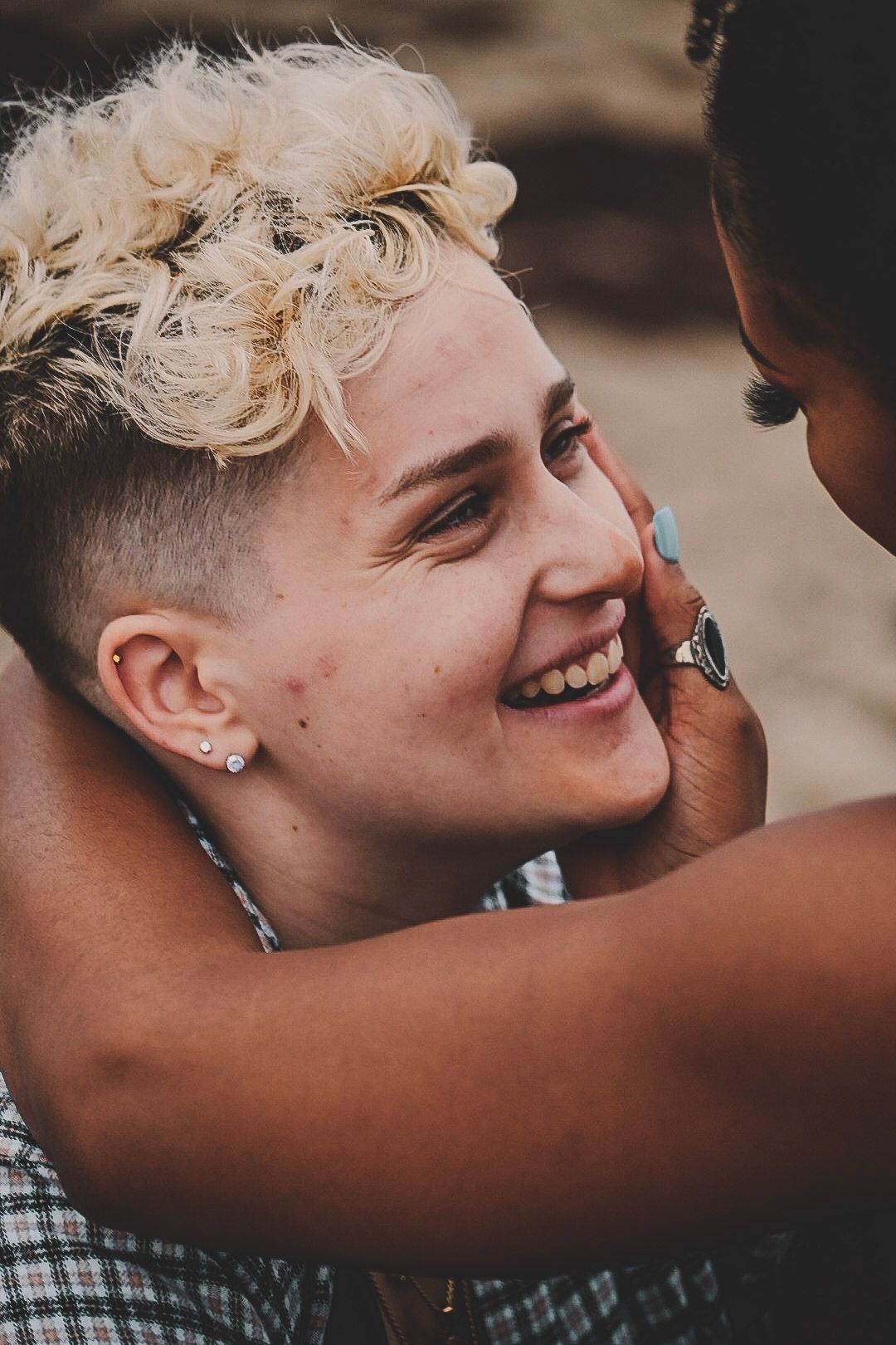 A smiling person with blonde curly undercut hair being tenderly touched on the face by another person outdoors.