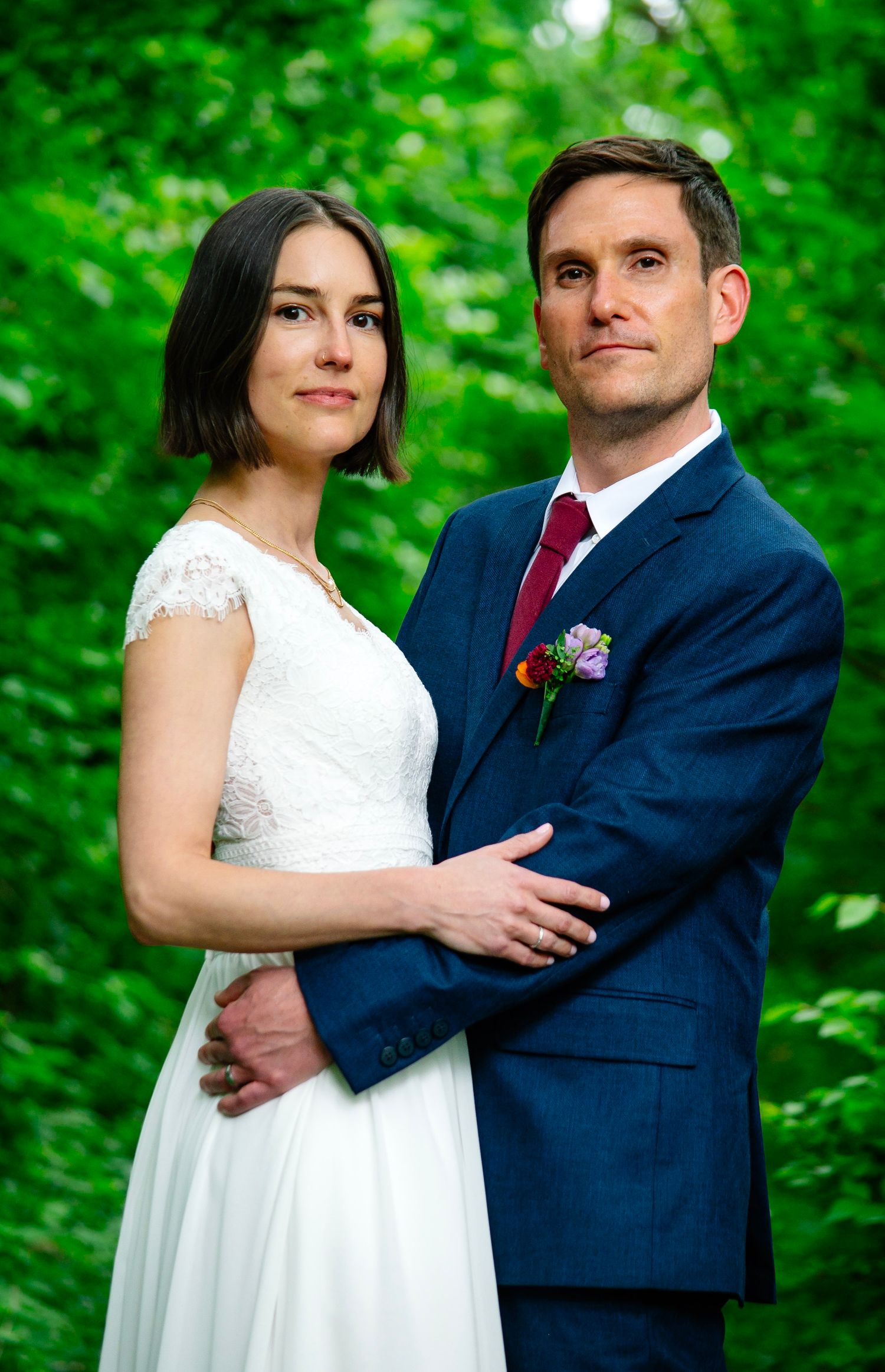 Bride in white lace dress and groom in navy suit with red tie pose together outdoors amid lush green foliage.
