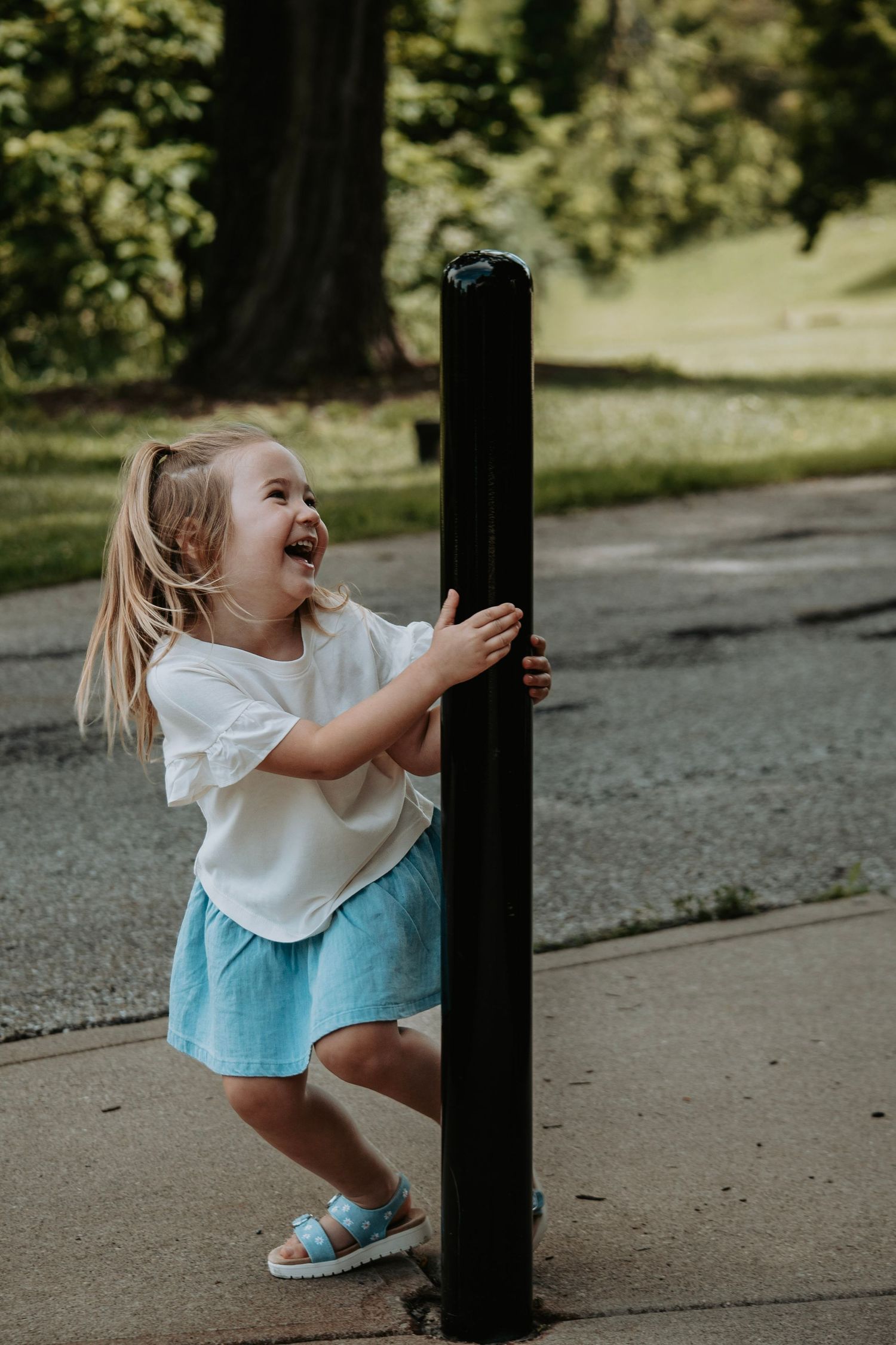 A joyful toddler girl in a white top and blue skirt laughs while holding a black bollard outdoors.