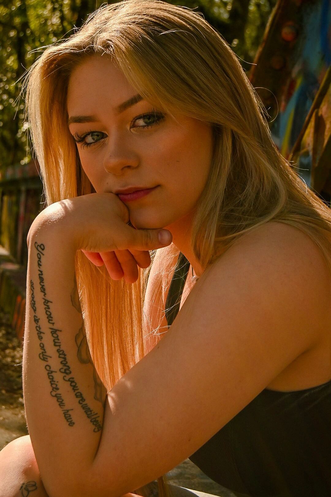 Blonde woman with arm tattoo posing outdoors in warm sunlight, wearing a black top.
