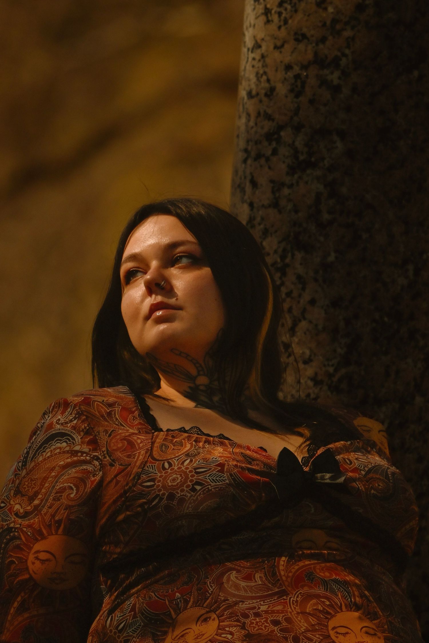 Woman in paisley dress leaning against stone column in warm golden light, gazing upward thoughtfully.