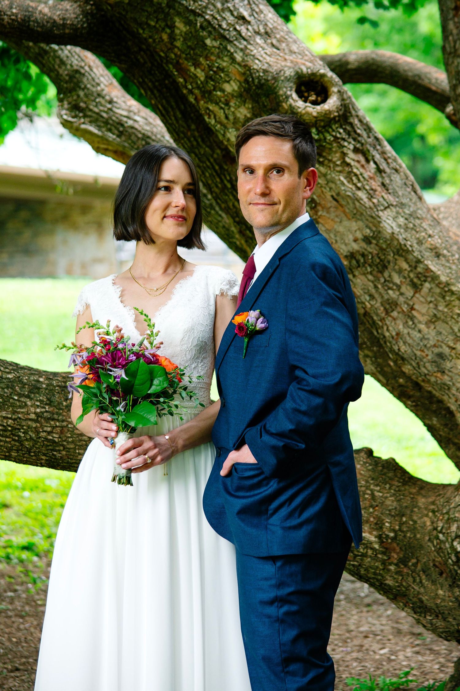Bride and groom posing under a large tree, she holds a colorful bouquet in a white dress, he wears a navy suit.