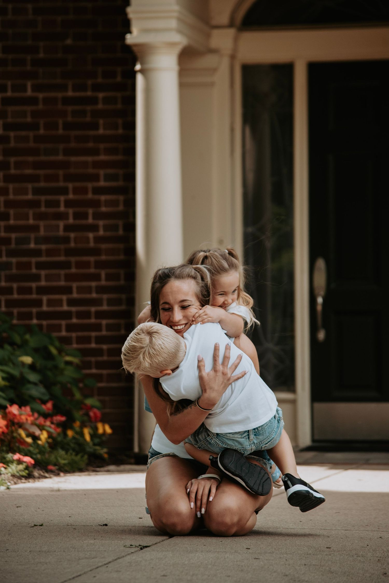 A smiling woman kneeling on a driveway hugging two young children in front of a brick house.