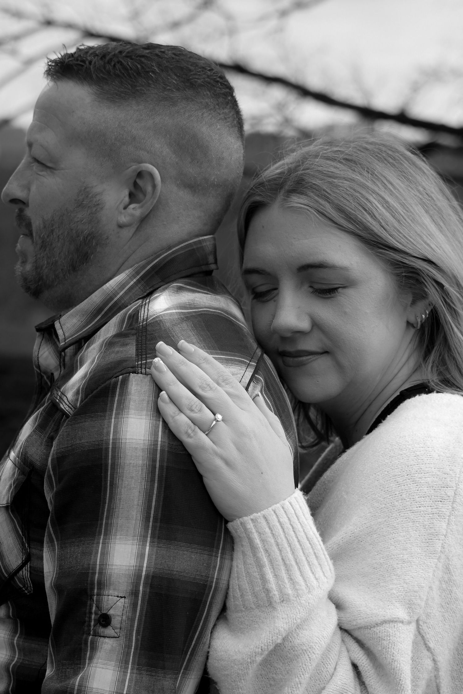 Black and white photo of a couple embracing, woman resting head on mans shoulder showing engagement ring.