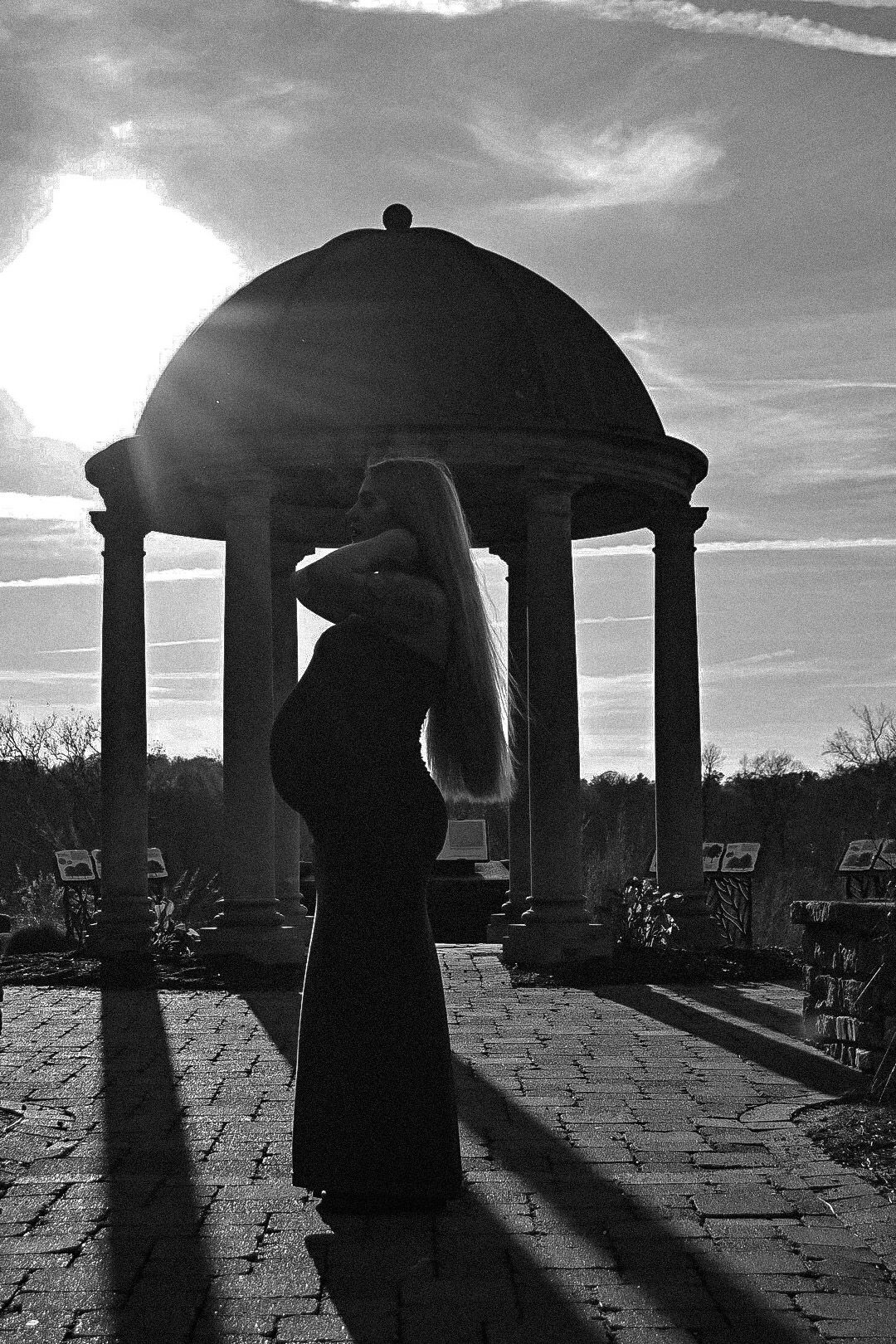 Silhouette of a pregnant figure standing under a domed gazebo at sunset casting dramatic shadows.
