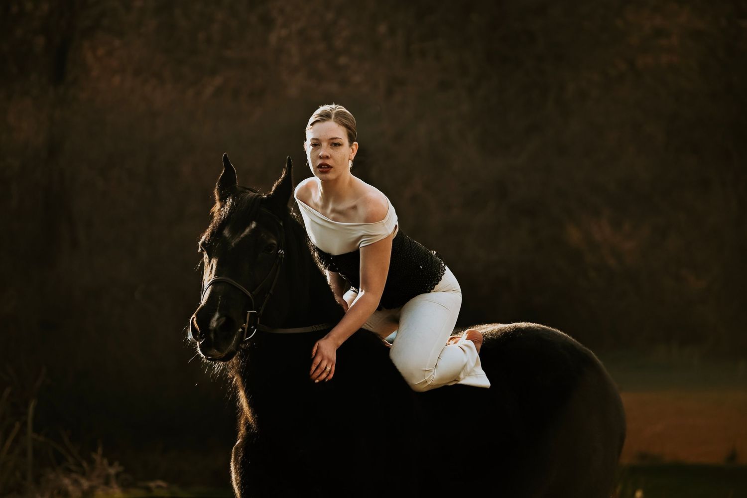 Young woman in white off-shoulder dress riding a dark horse in a moody outdoor setting.