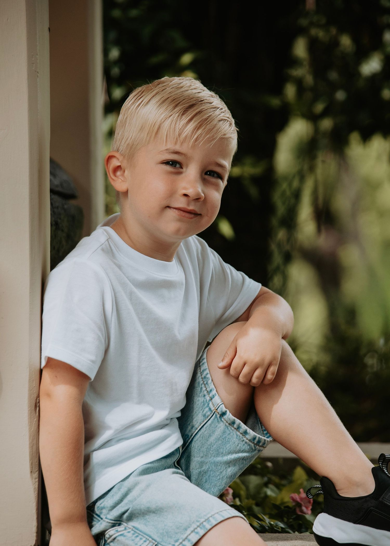 Adorable blonde boy in white t-shirt and denim shorts sits relaxed outdoors with a gentle smile.