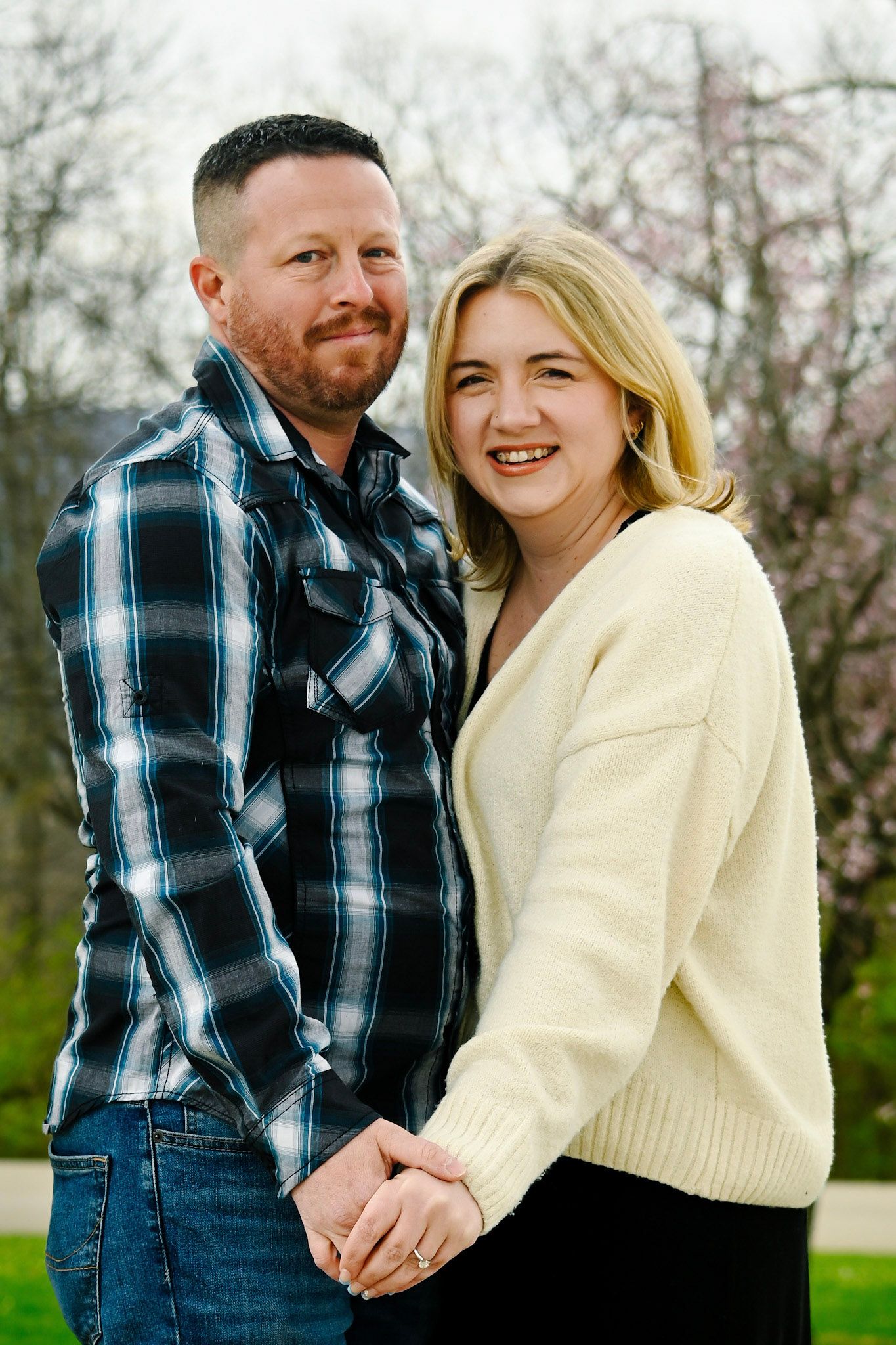 A smiling couple holds hands outdoors, man in plaid shirt and woman in cream sweater, with blooming trees in background.